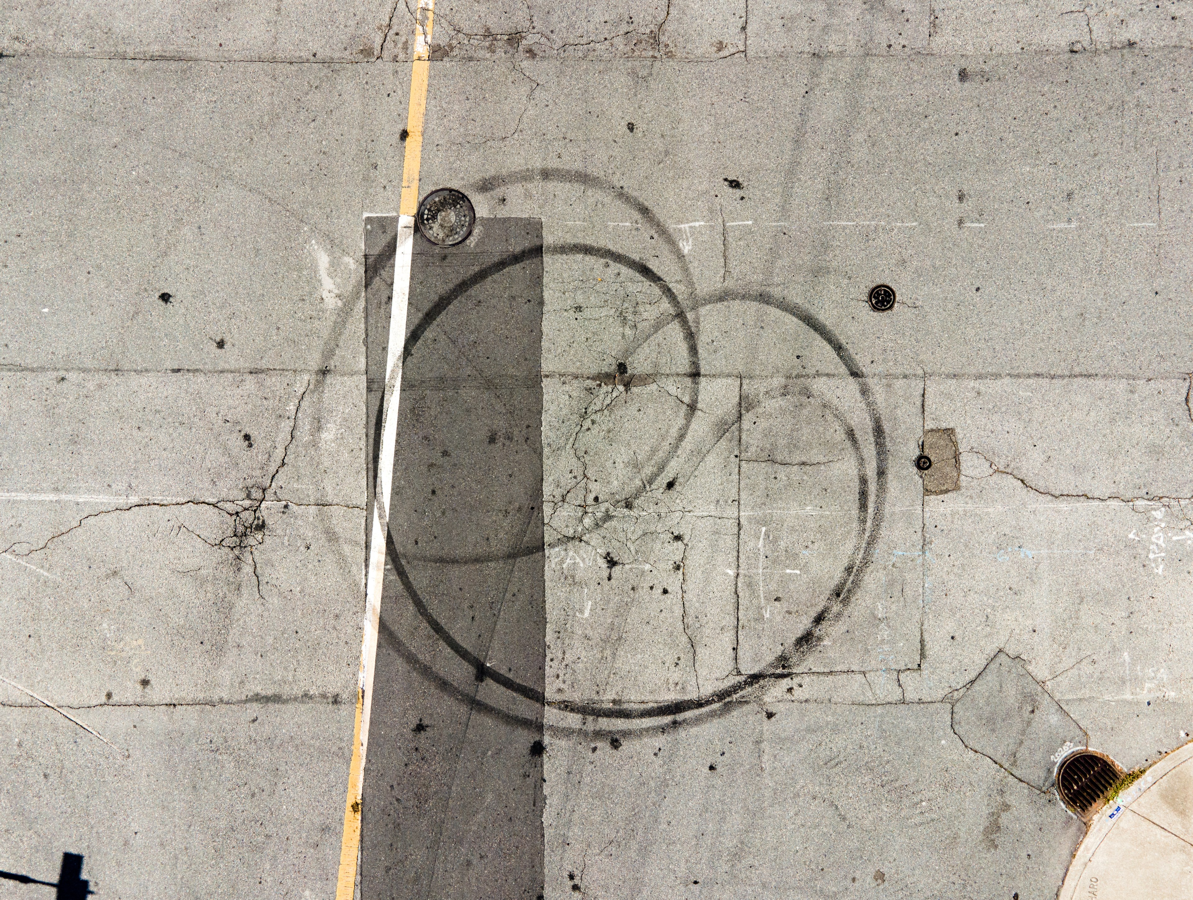 Aerial view of a cracked asphalt surface with circular tire marks, a yellow line, and two manholes, one on the left and another on the right near a drain.