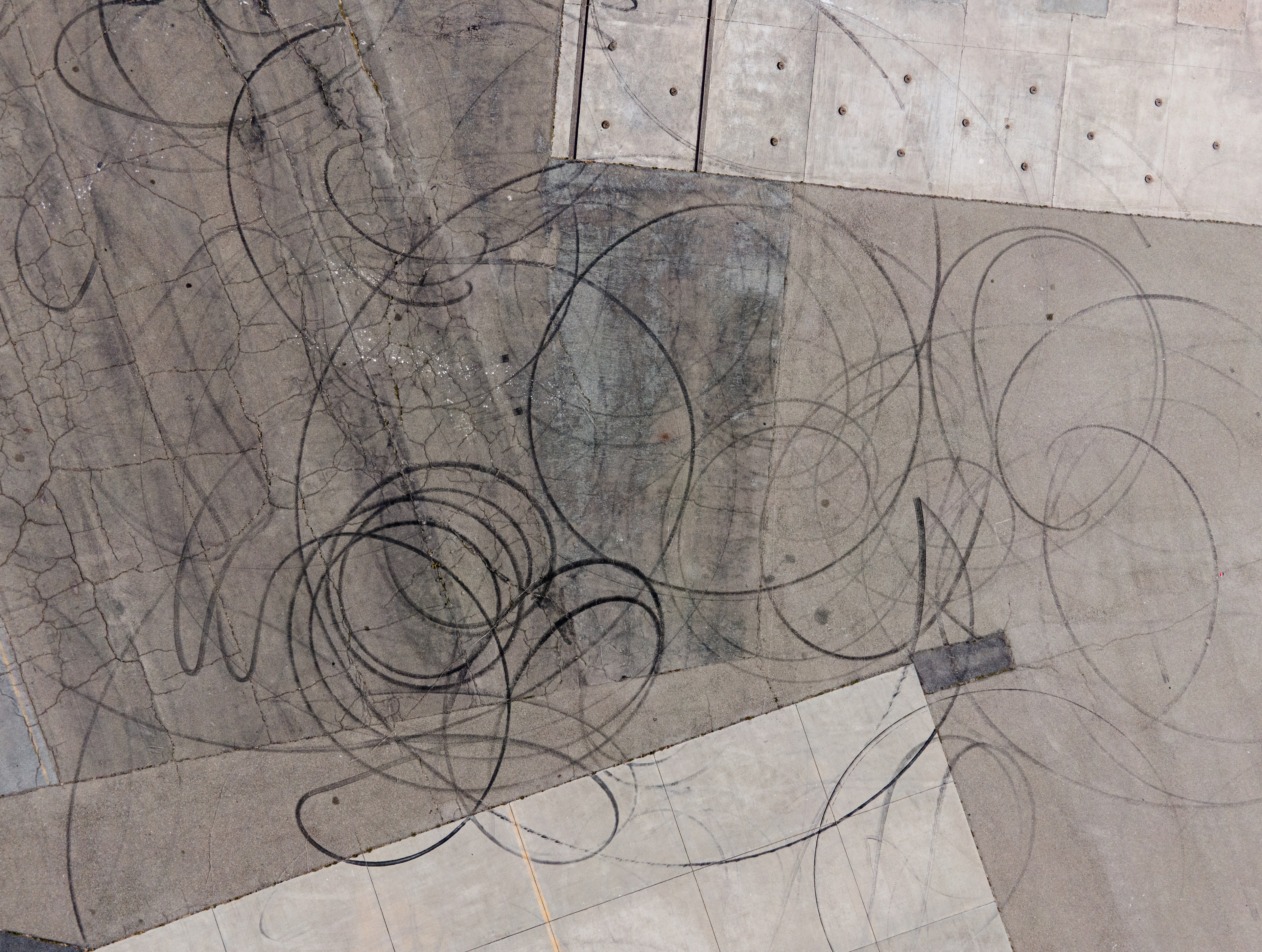The image shows an aerial view of a concrete surface covered with circular tire skid marks, creating a chaotic pattern. The ground appears weathered and cracked.