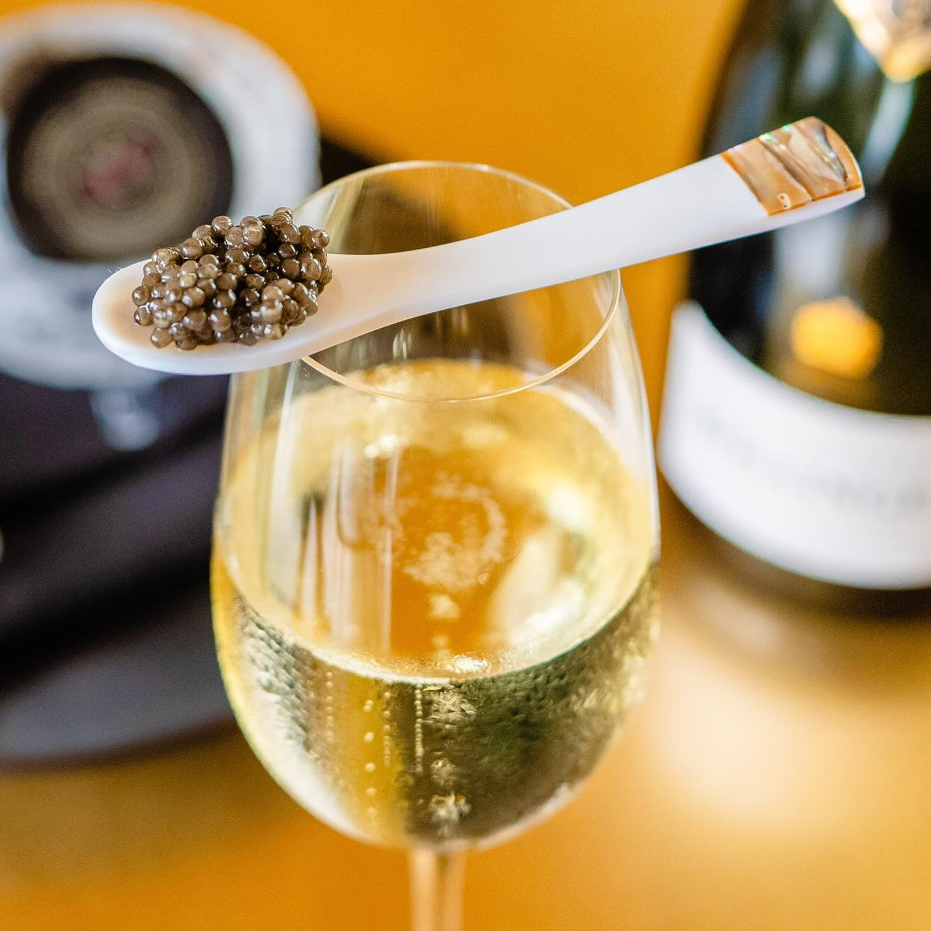 A glass of champagne is topped with a white spoon holding black caviar. In the background, there's a blurred bottle and container.