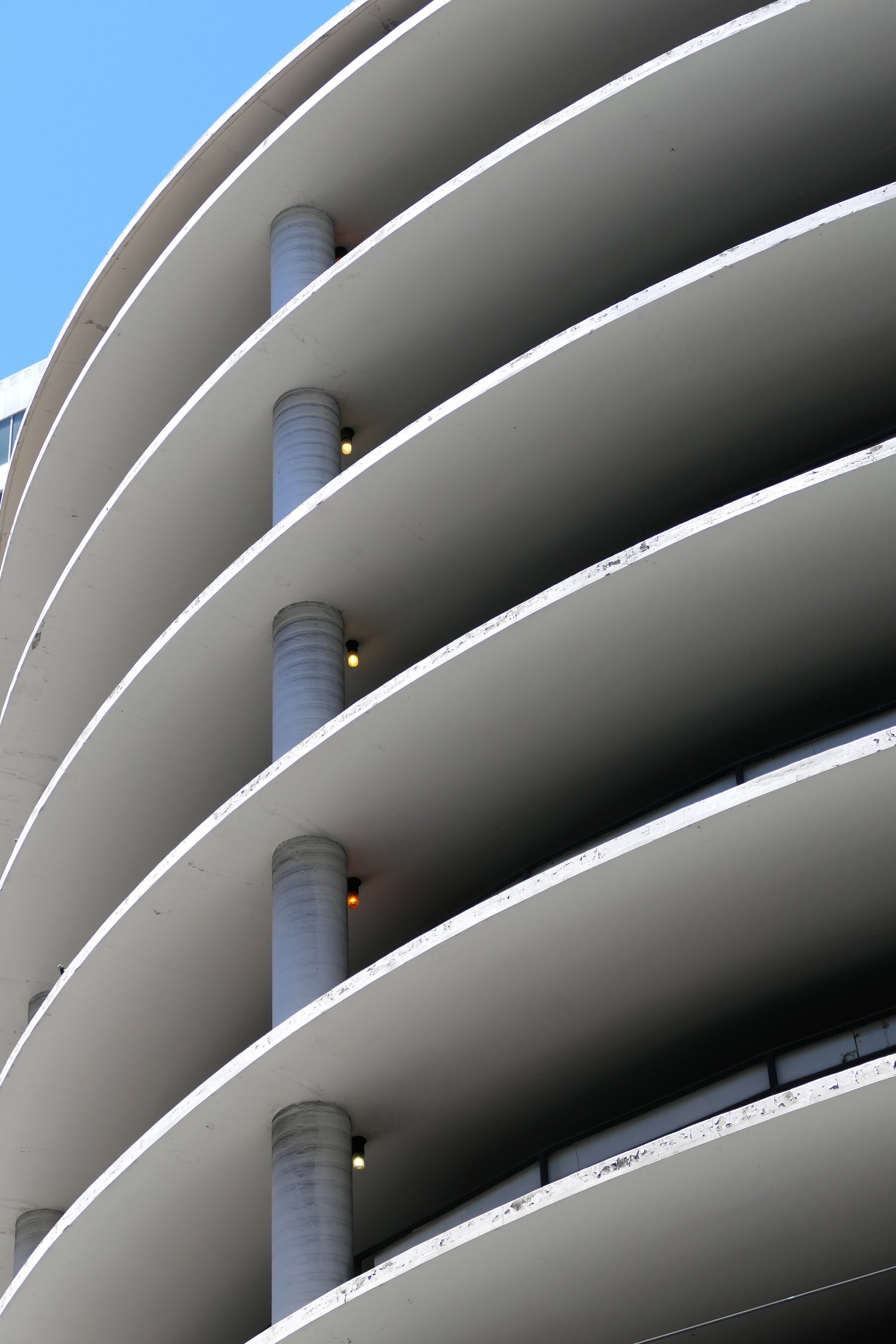 The image shows a modern building with curved, layered floors and circular columns, against a clear blue sky. Small lights are attached near each column.