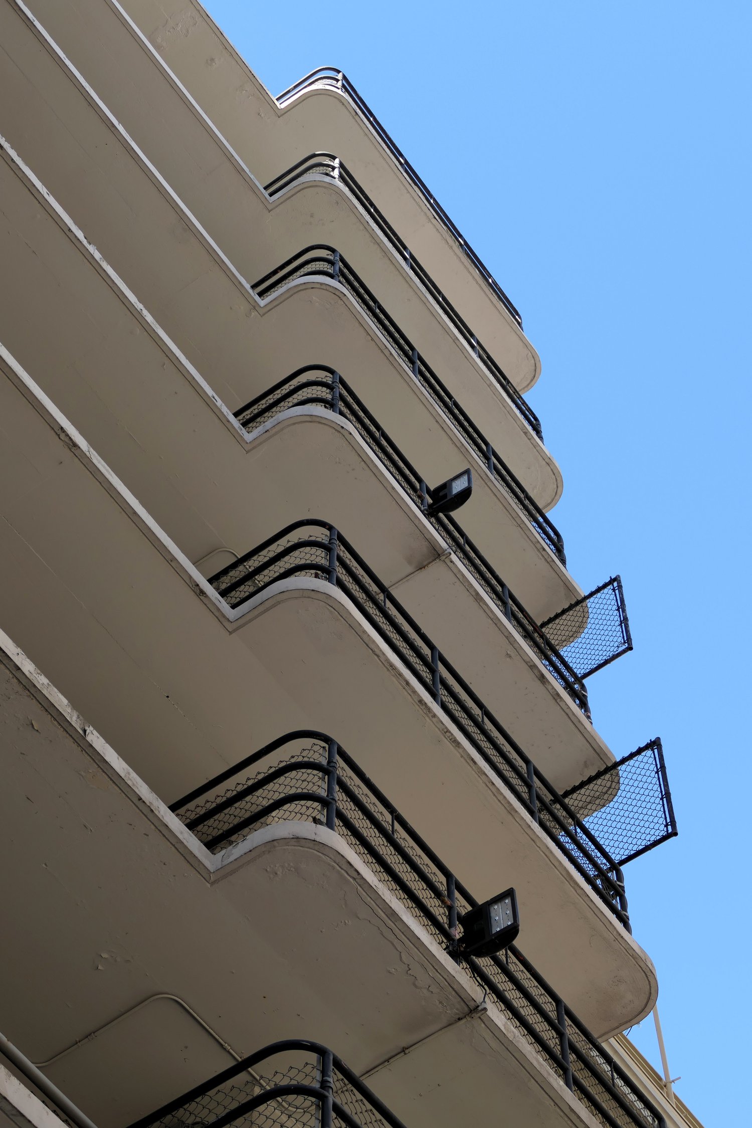 The image shows several levels of a building with curved balconies, featuring black railings and safety nets, set against a clear blue sky.