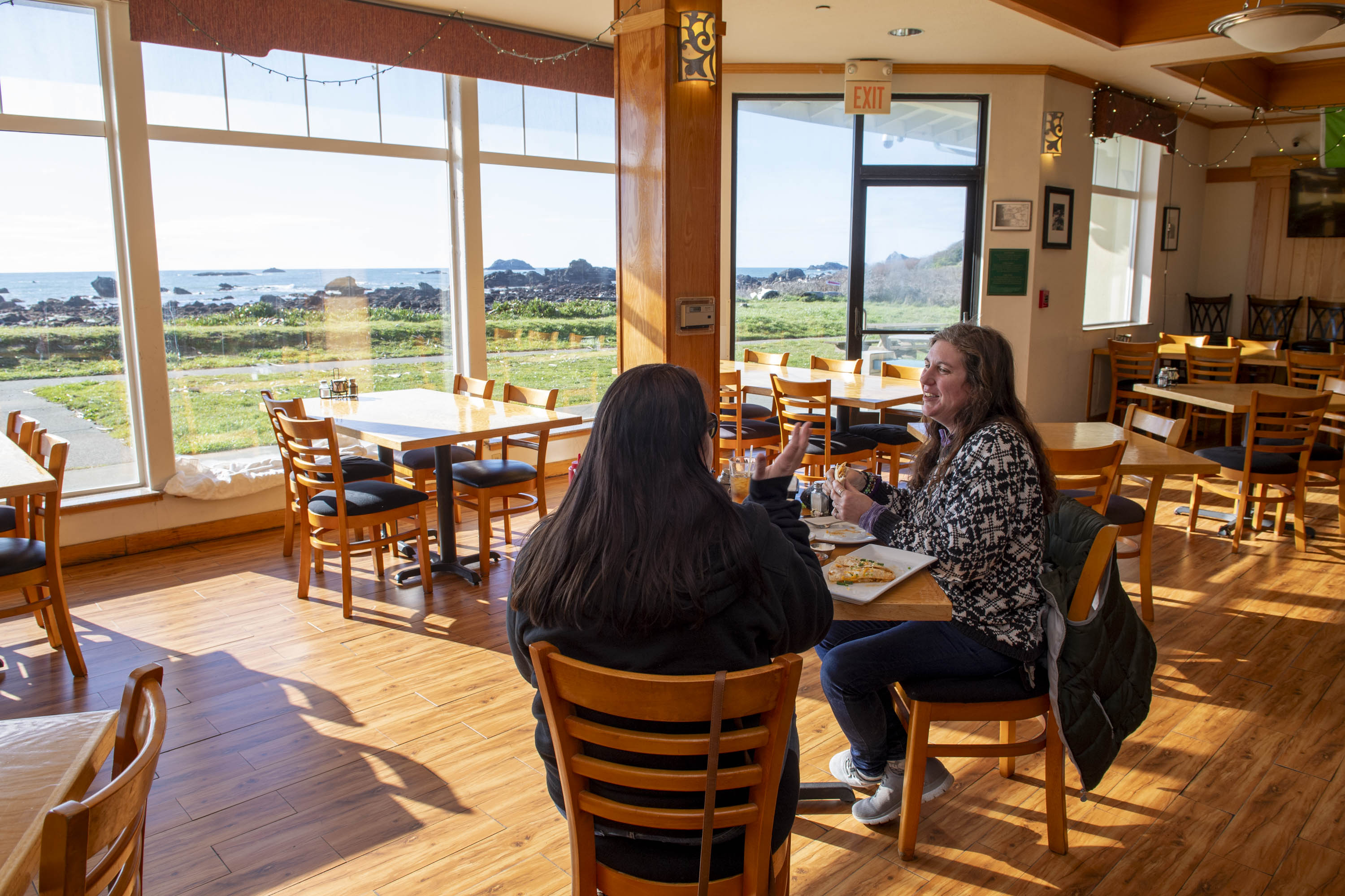 Two people sit at a wooden table in a sunlit restaurant with large windows, offering a view of green grass and the ocean. They appear to be chatting and eating.
