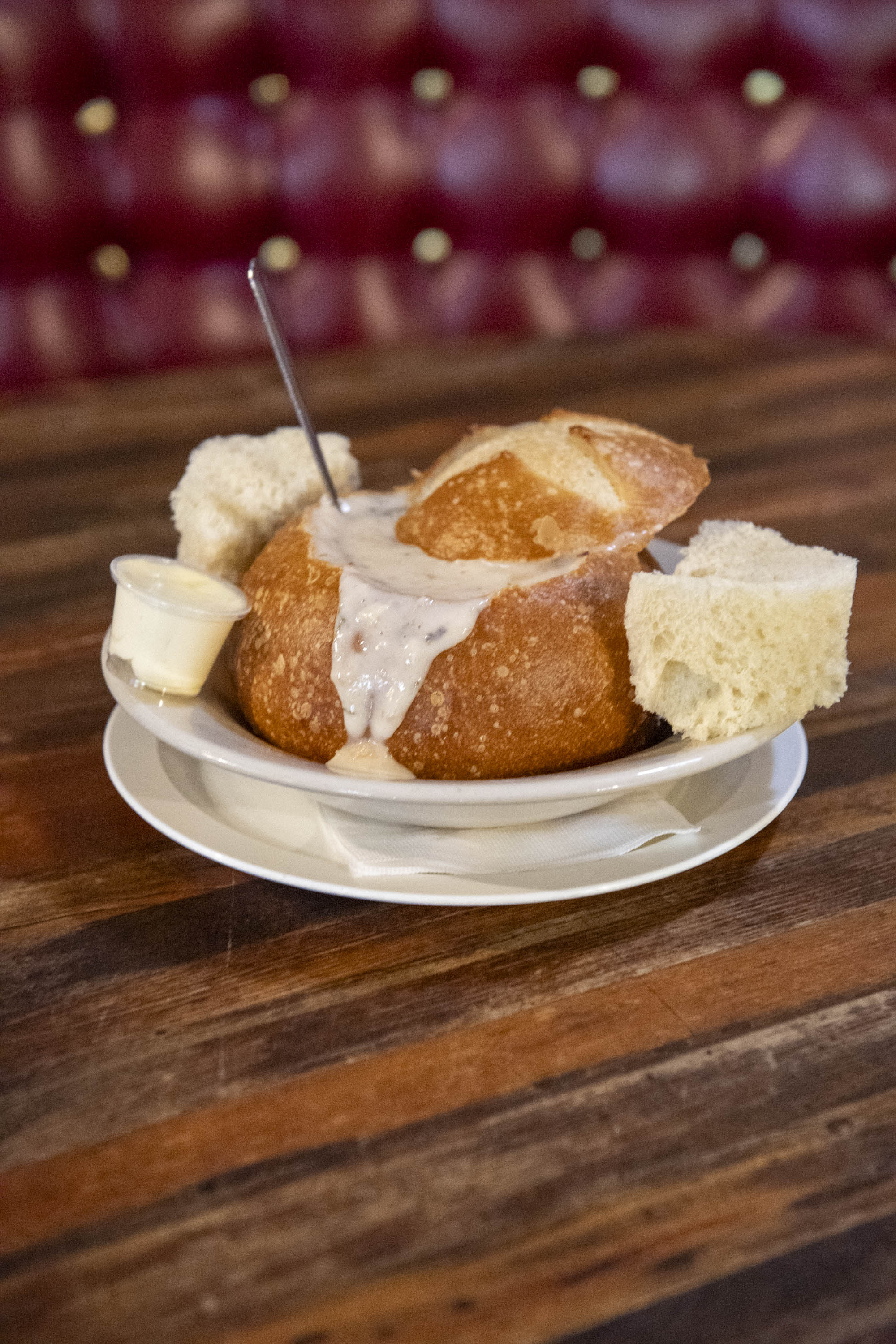 A bread bowl filled with creamy soup sits on a plate with bread pieces and a small container of butter. A spoon is placed in the soup.
