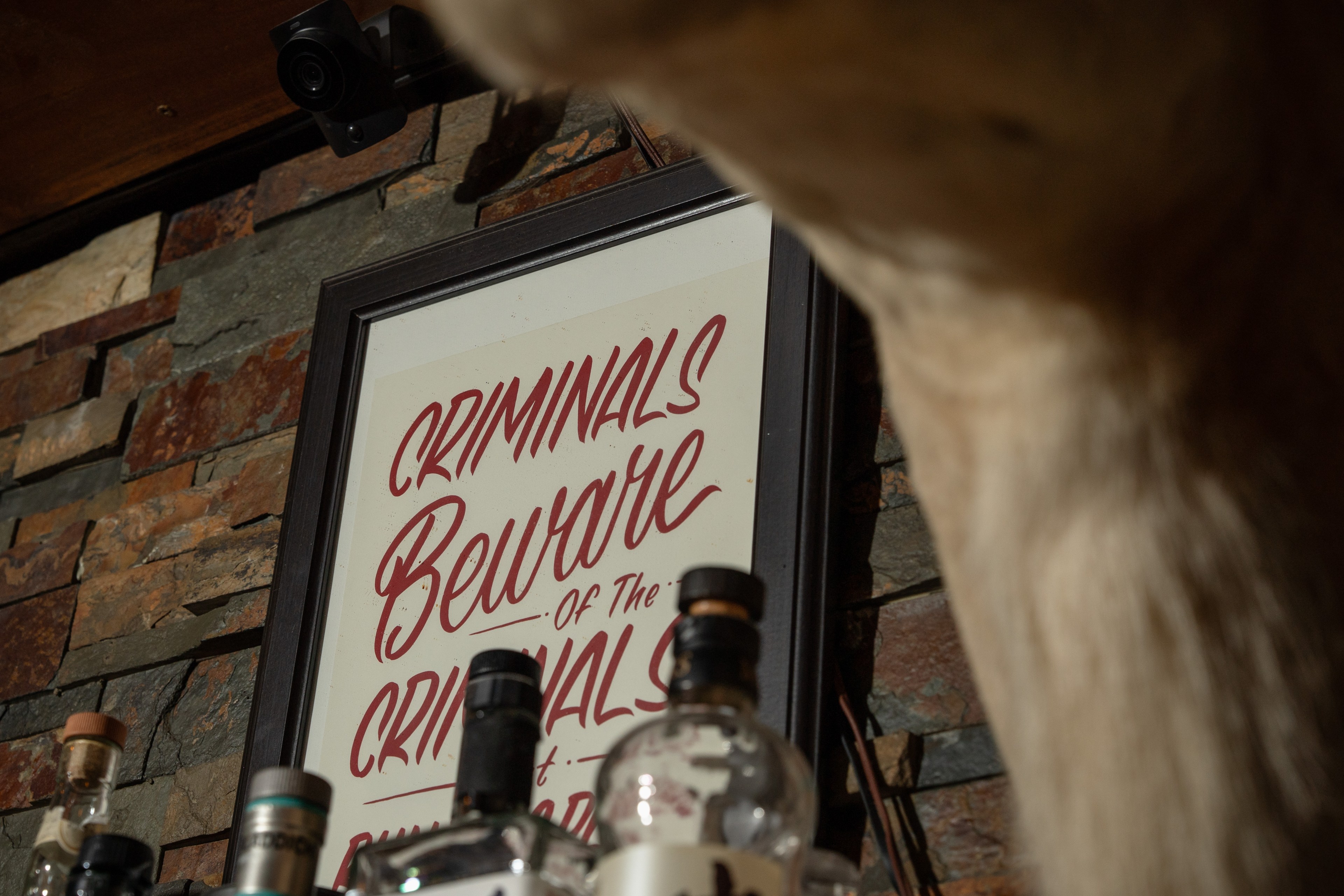 The image shows a framed sign reading &quot;CRIMINALS BEWARE&quot; on a stone wall, partially blocked by bottles and a furry animal, possibly a dog's back.