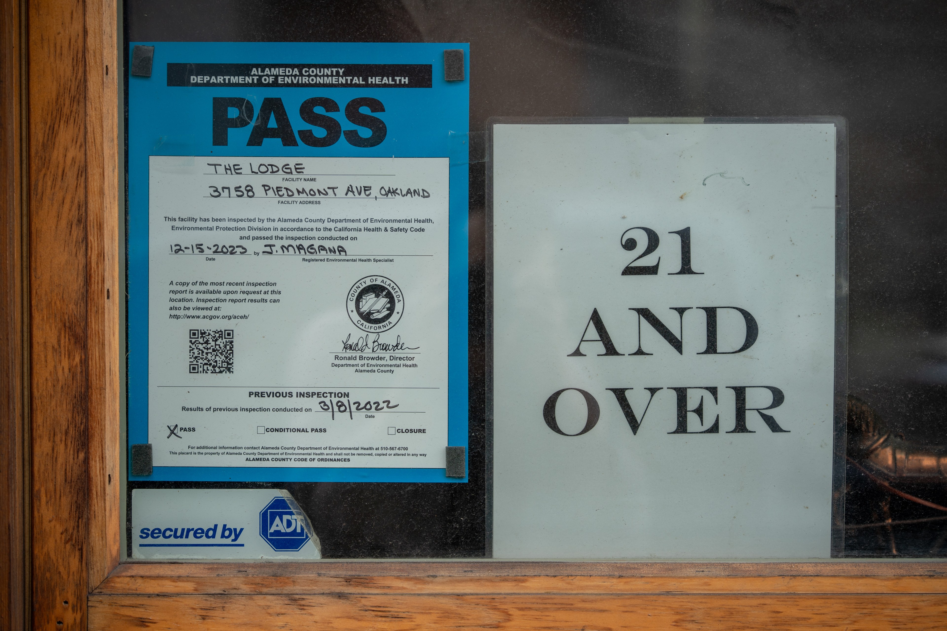 A blue health inspection pass from Alameda County is displayed next to a sign stating “21 AND OVER.” An ADT security decal is also visible.