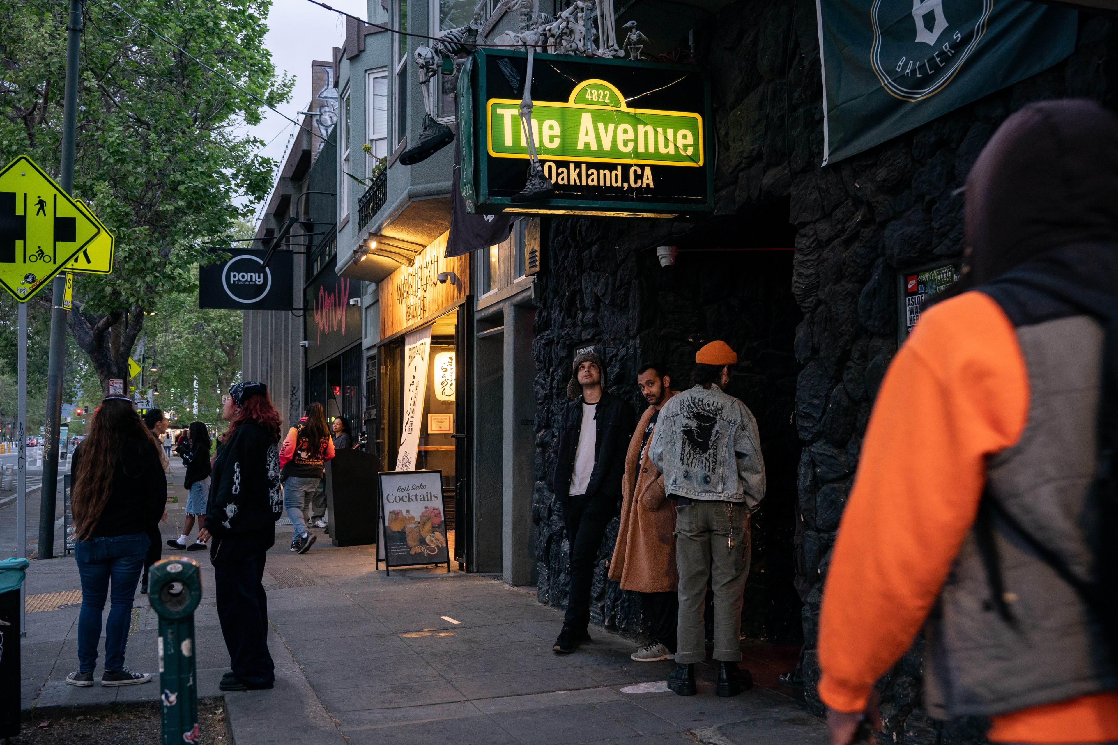 The image shows a lively street in Oakland, California, with people gathered outside &quot;The Avenue&quot; bar. A sidewalk sign advertises cocktails, and there's a neon pedestrian sign.