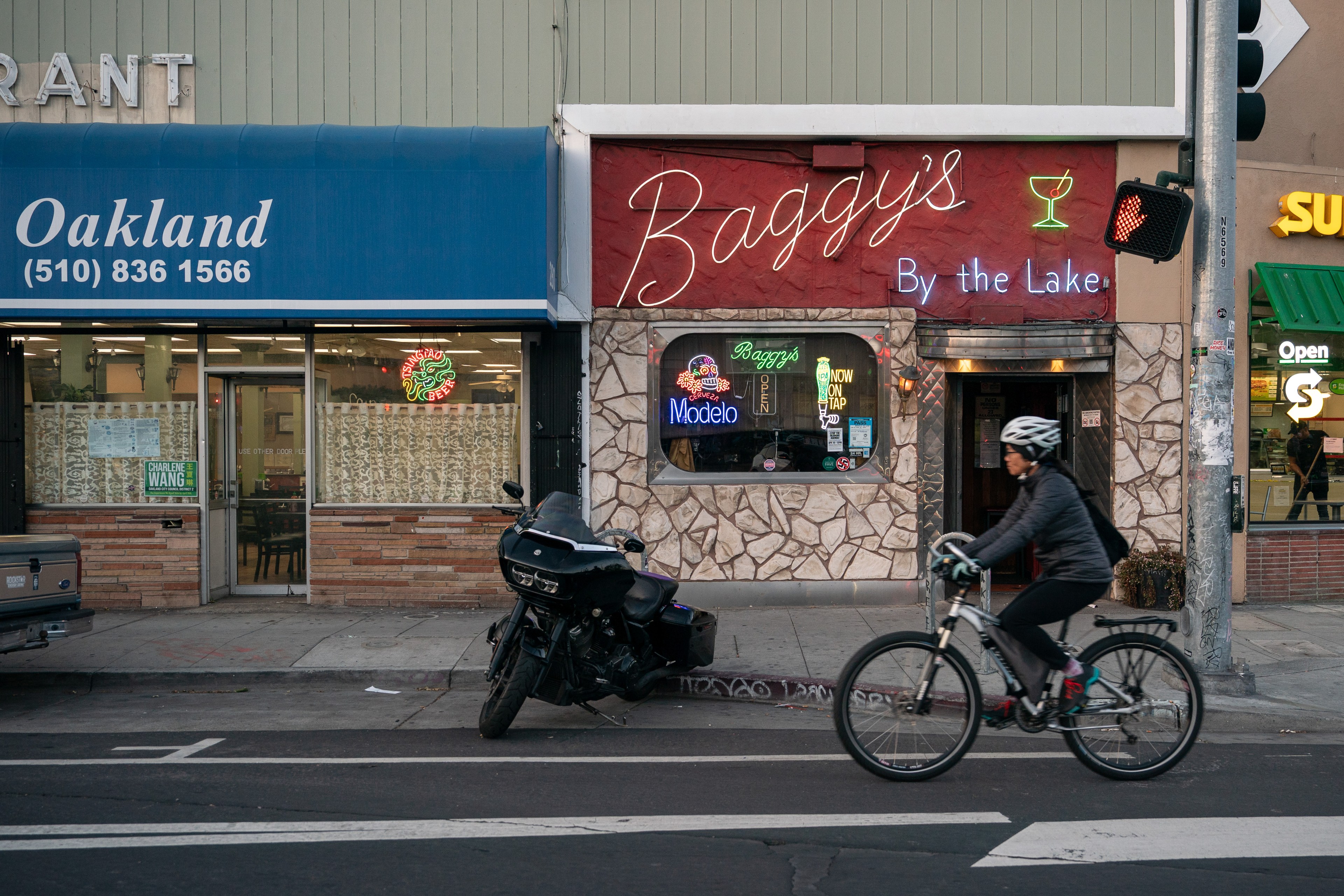 A cyclist rides past a row of storefronts including Baggy's Bar and an Oakland restaurant. A motorcycle is parked on the sidewalk beside the bar.