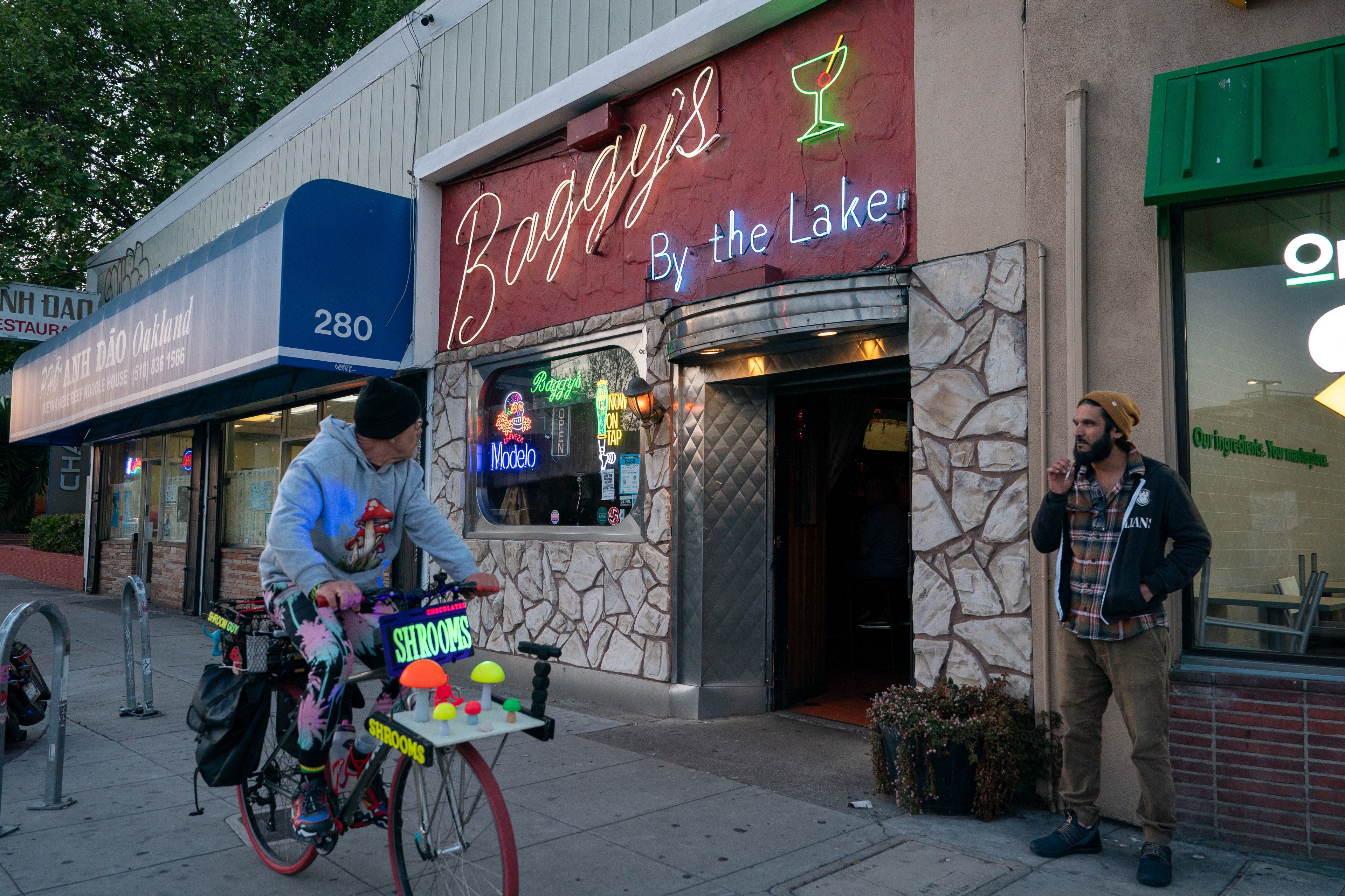 A person rides a bike decorated with colorful mushroom signs past a bar called "Baggy's By the Lake." Another person stands by the entrance, smoking.