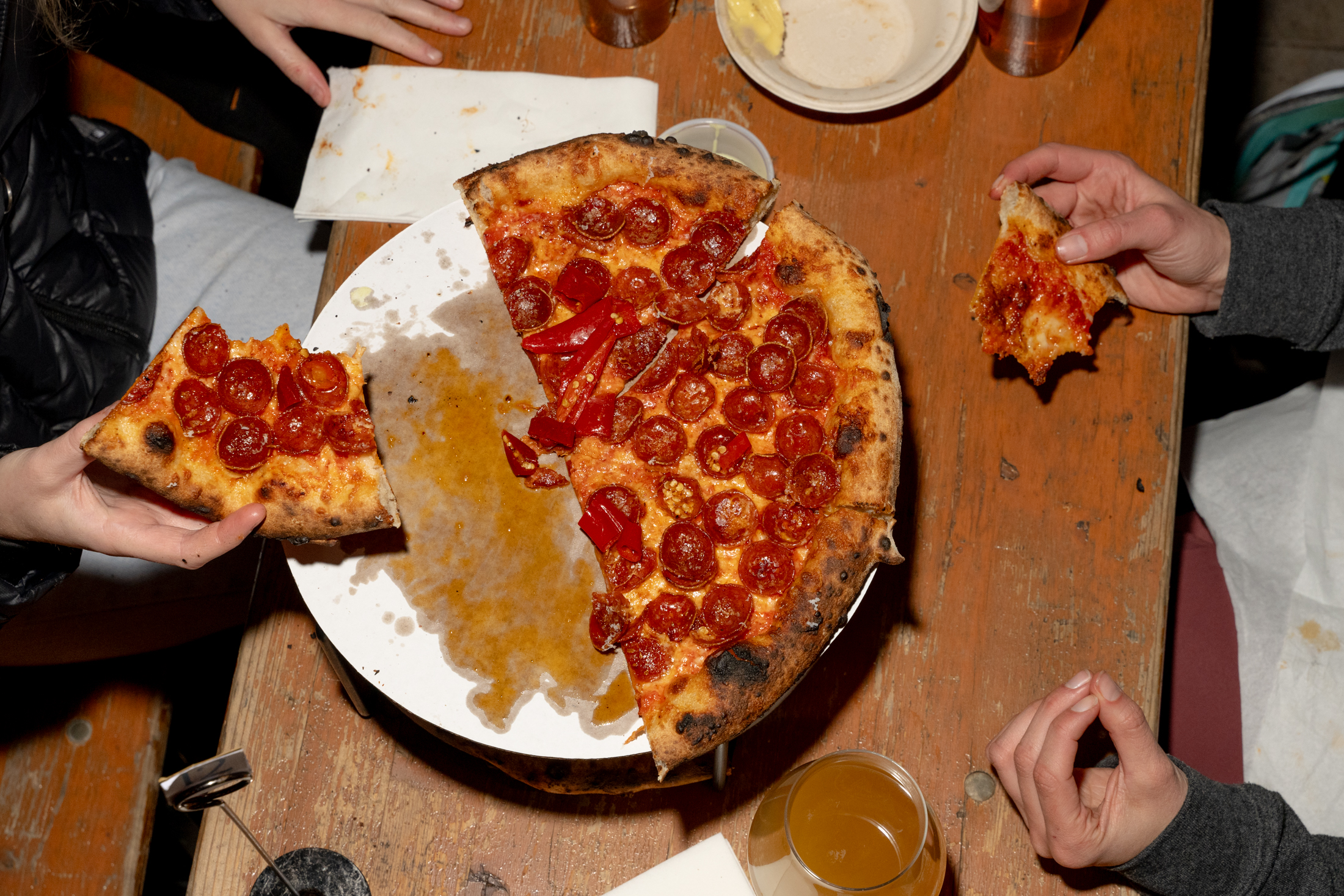 A partially eaten pepperoni pizza sits on a white plate on a wooden table. Three people are holding slices, and there's a drink nearby.