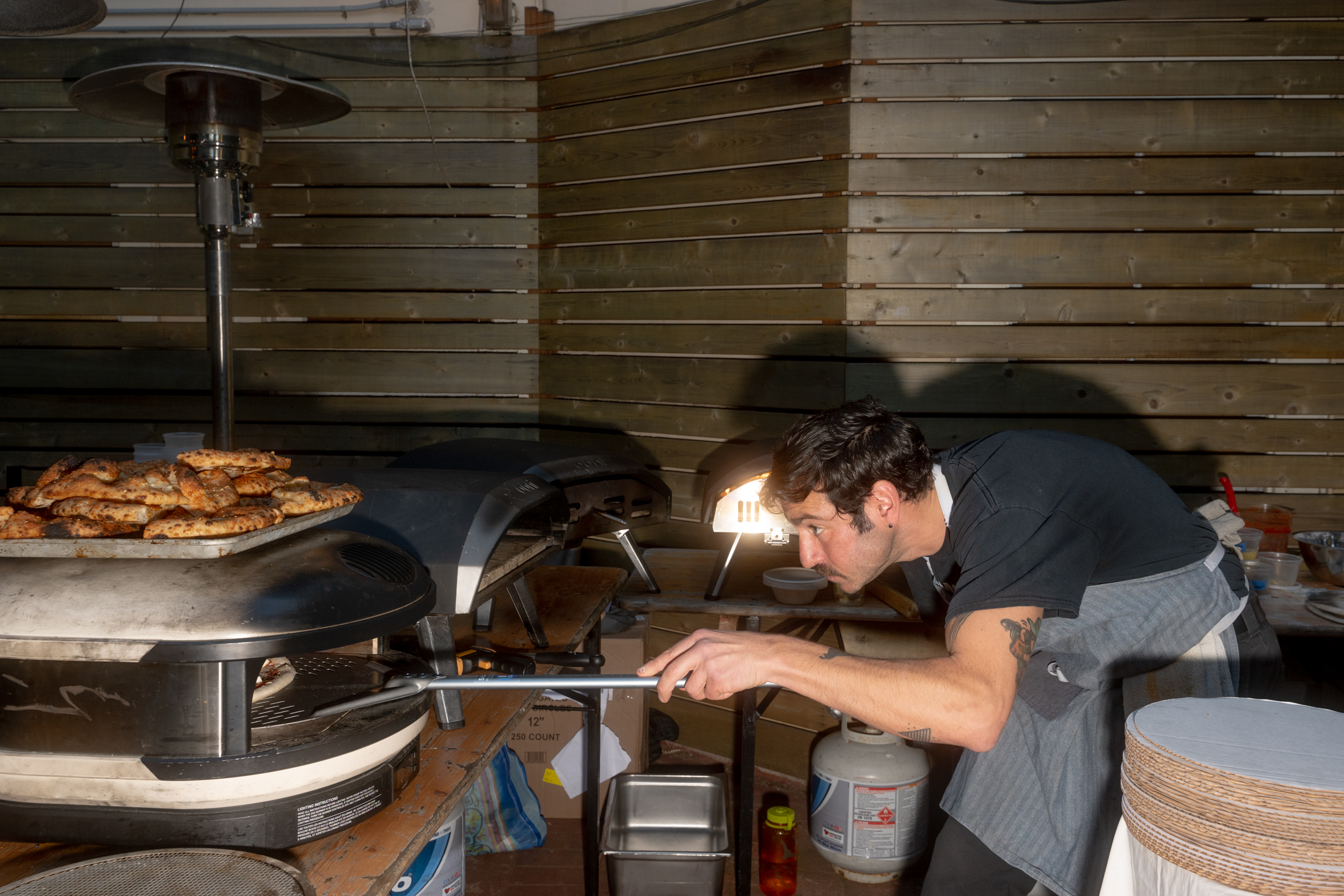 A person in a black shirt and apron is focused on baking pizza in an outdoor oven. A tray of cooked pizzas is nearby, and a wooden wall forms the background.
