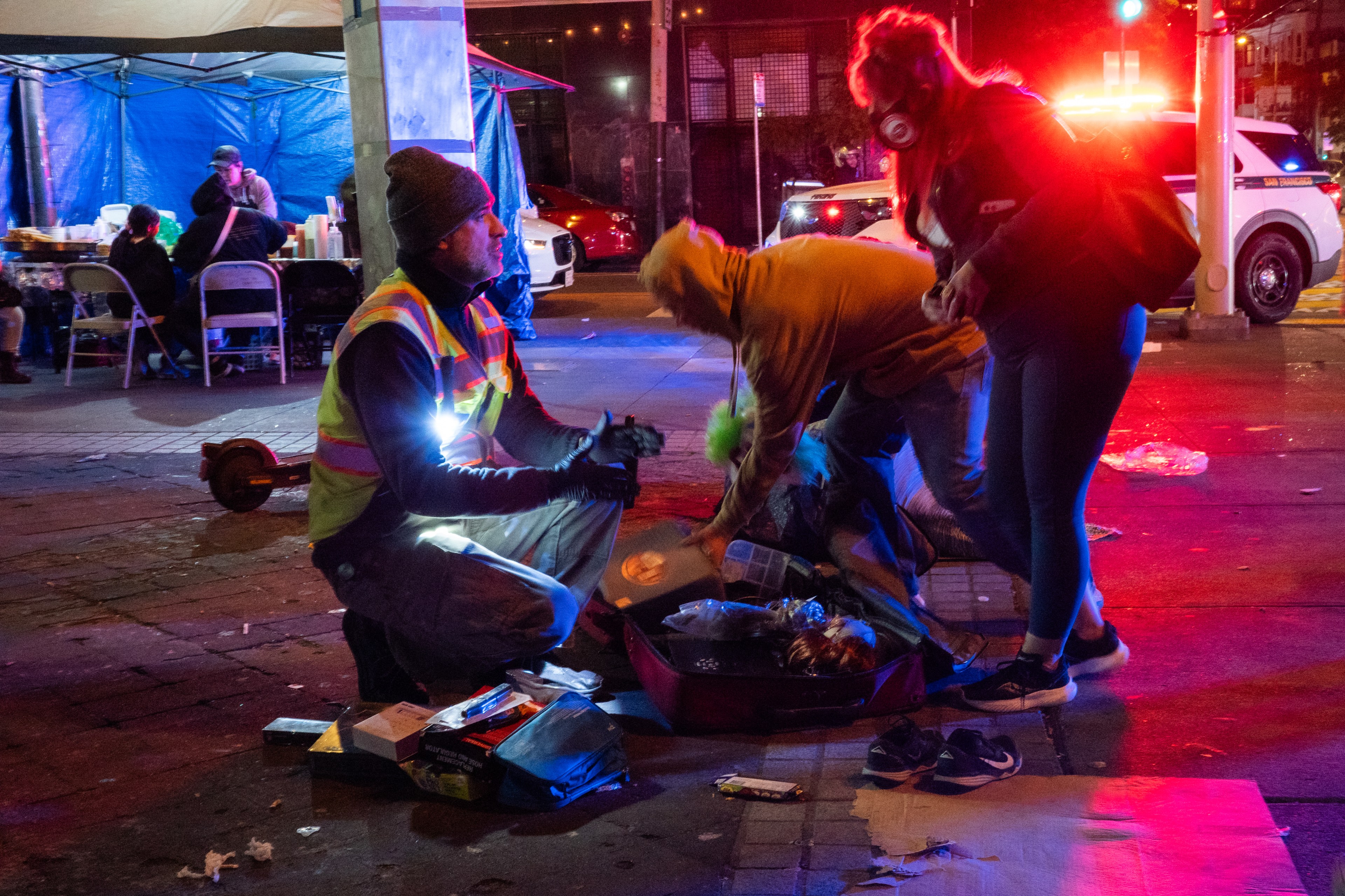 Several people interact on a dimly lit street in front of a tent with emergency lights flashing in the background. One person wears a reflective vest.