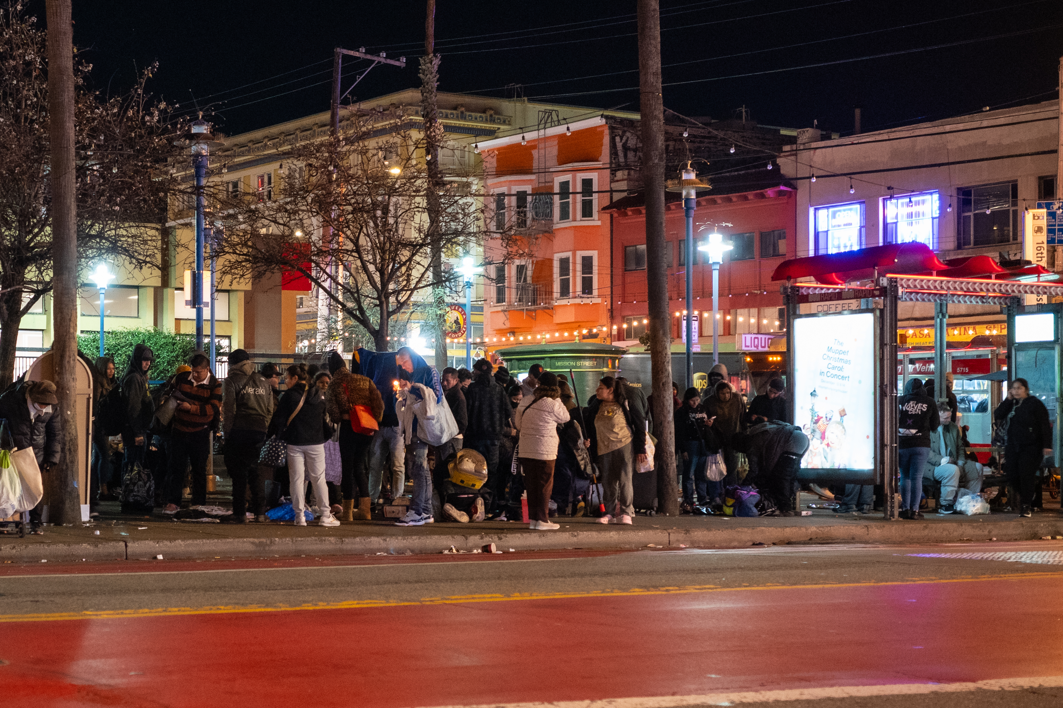 A large group of people gather at a bus stop on a city street at night. Buildings with bright lights and signs are visible in the background.