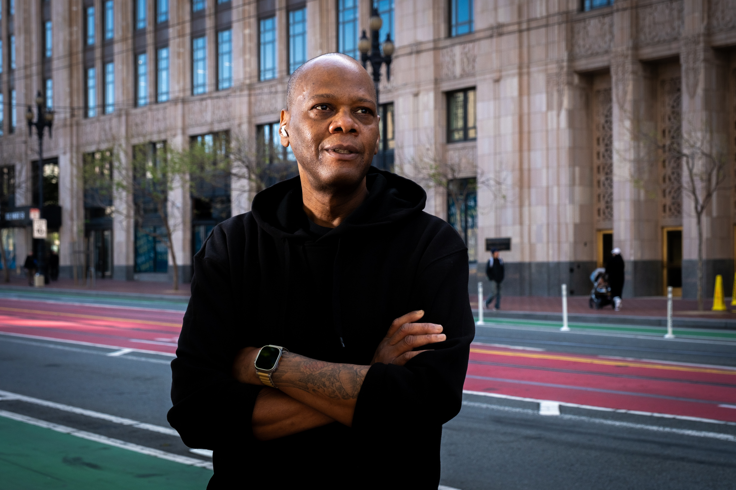 A man stands on Market street in a black hoodie with his arms crossed.