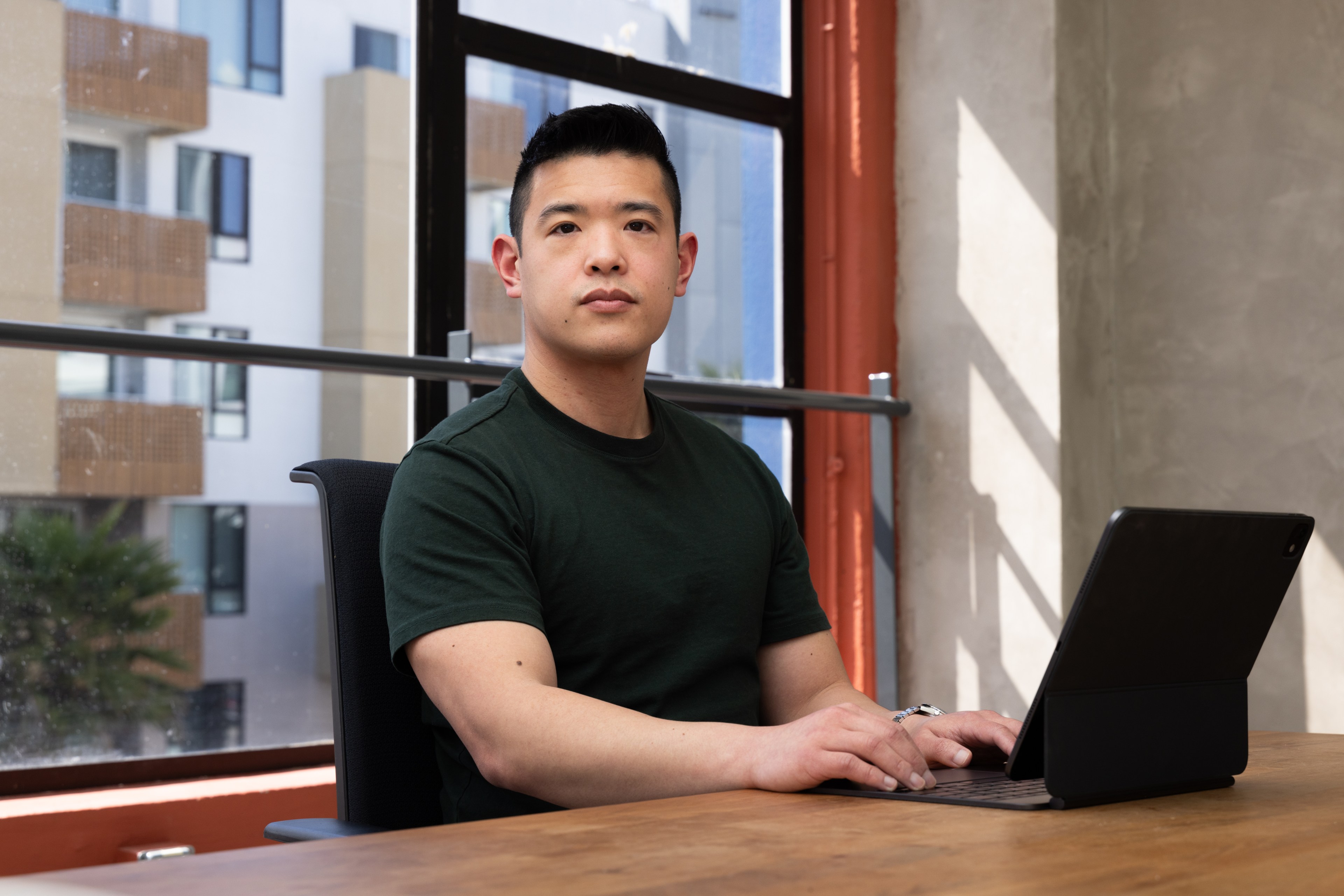 A person in a dark green shirt sits at a wooden table, typing on a laptop in front of a large window with a view of modern apartment buildings outside.