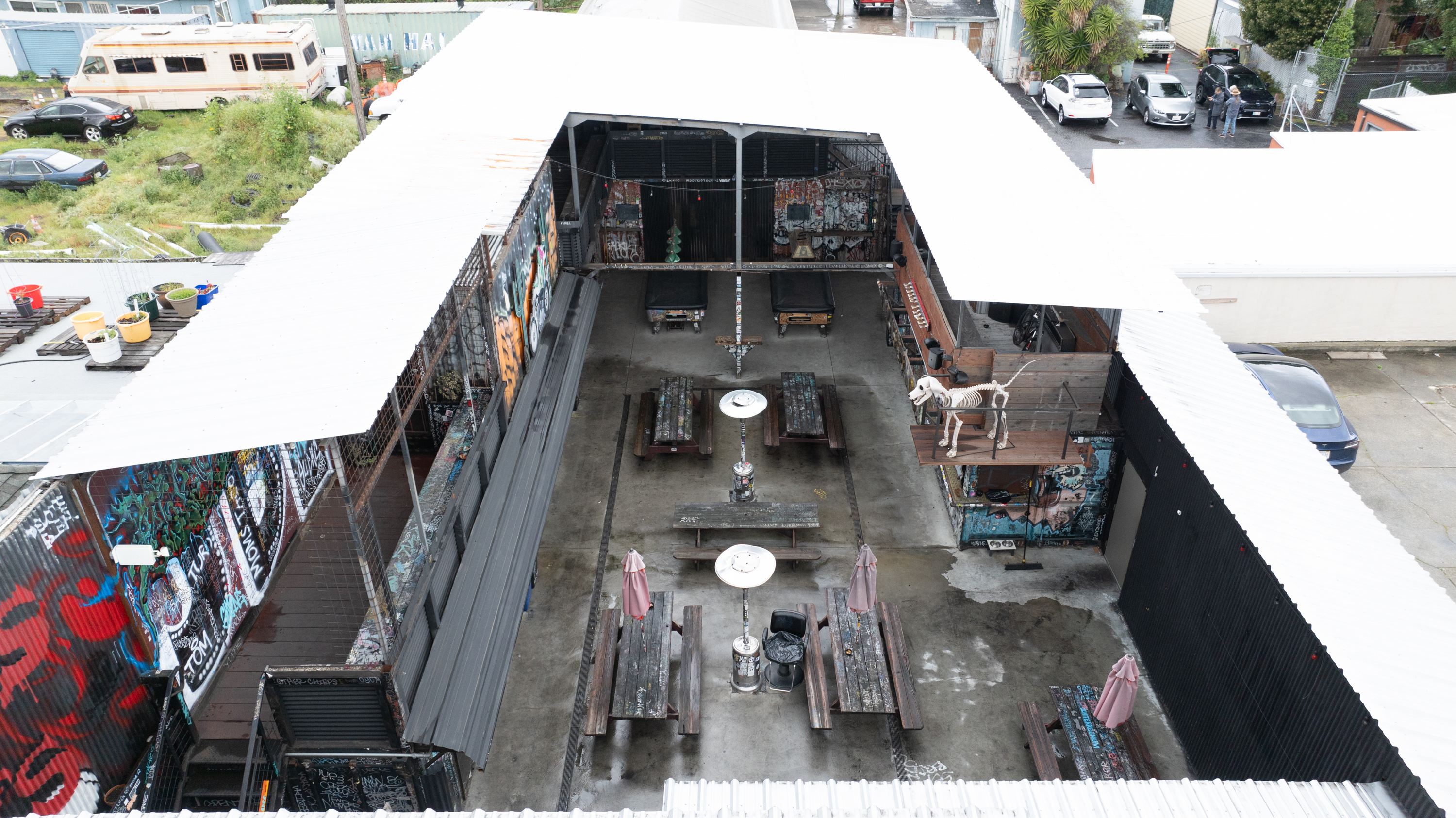 The image shows a U-shaped rooftop terrace with graffiti-covered walls and several picnic tables. A sculpture of a skeleton dog is on the right.