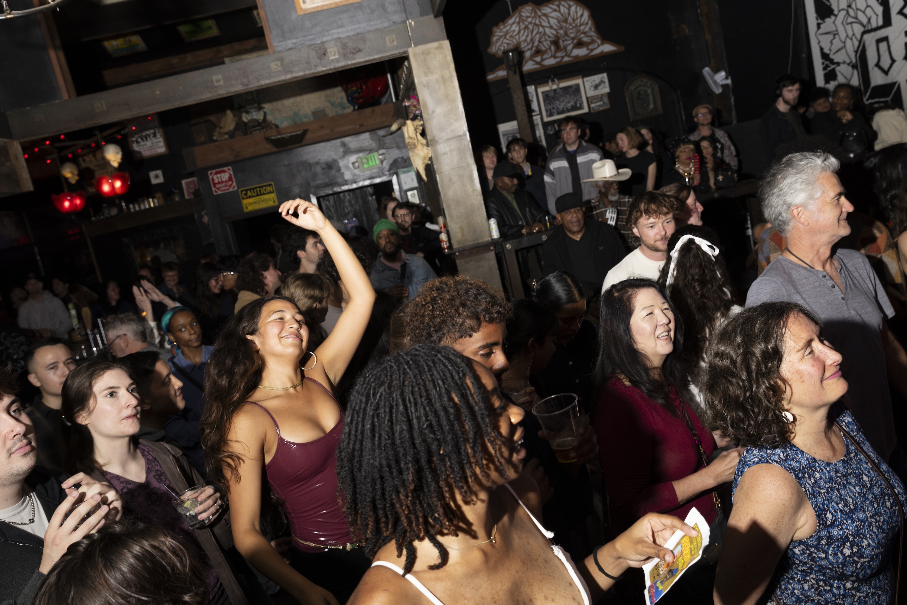 A lively crowd at a dimly lit venue, with people smiling, drinking, and dancing. Decor includes red lights and various wall art, creating a festive atmosphere.