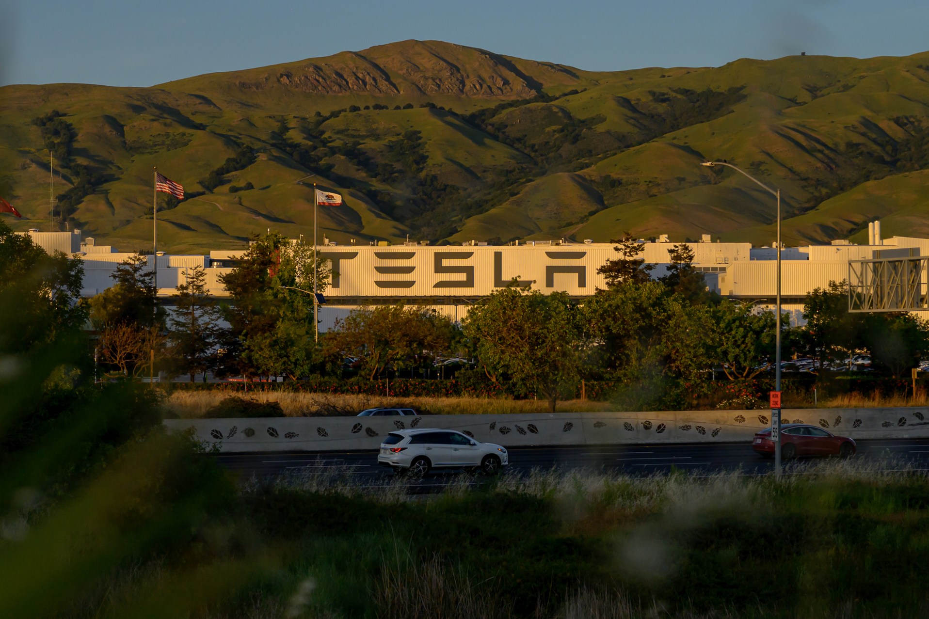 A white Tesla building is set against green hills, with flags flying above. Trees partially block the view, and cars travel on a road in the foreground.
