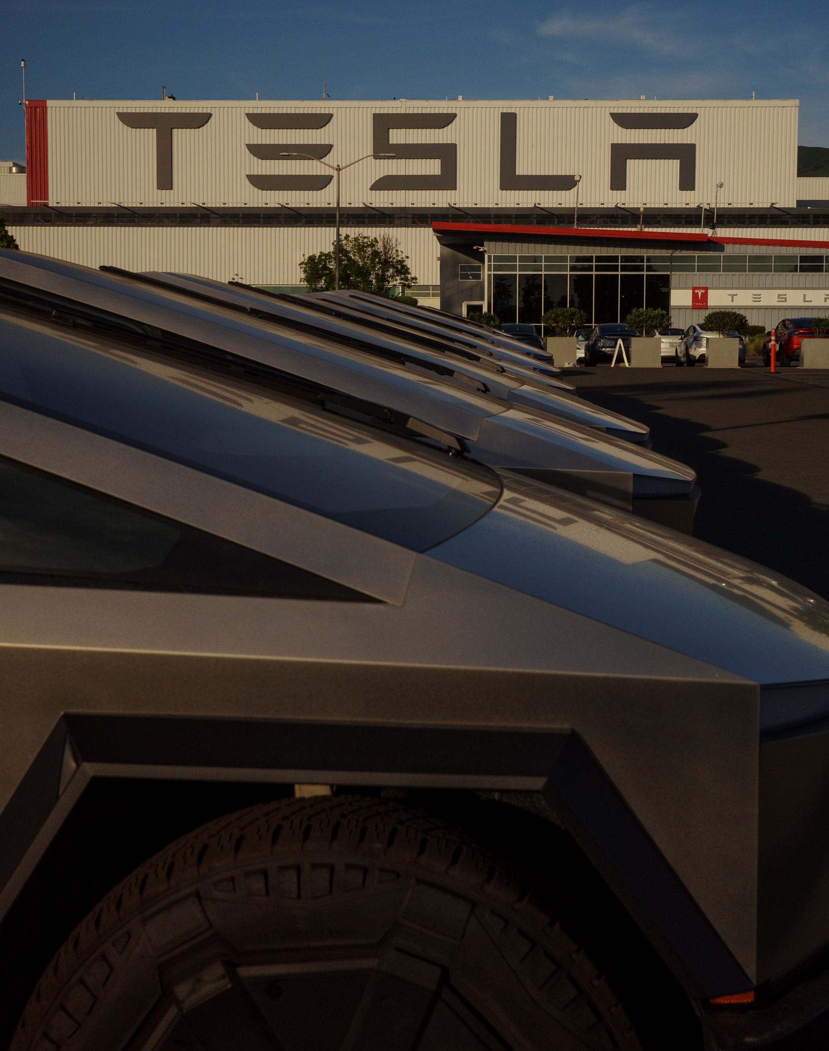 The image shows several sleek, angular trucks parked in front of a large building with &quot;TESLA&quot; displayed prominently in bold letters on the side.