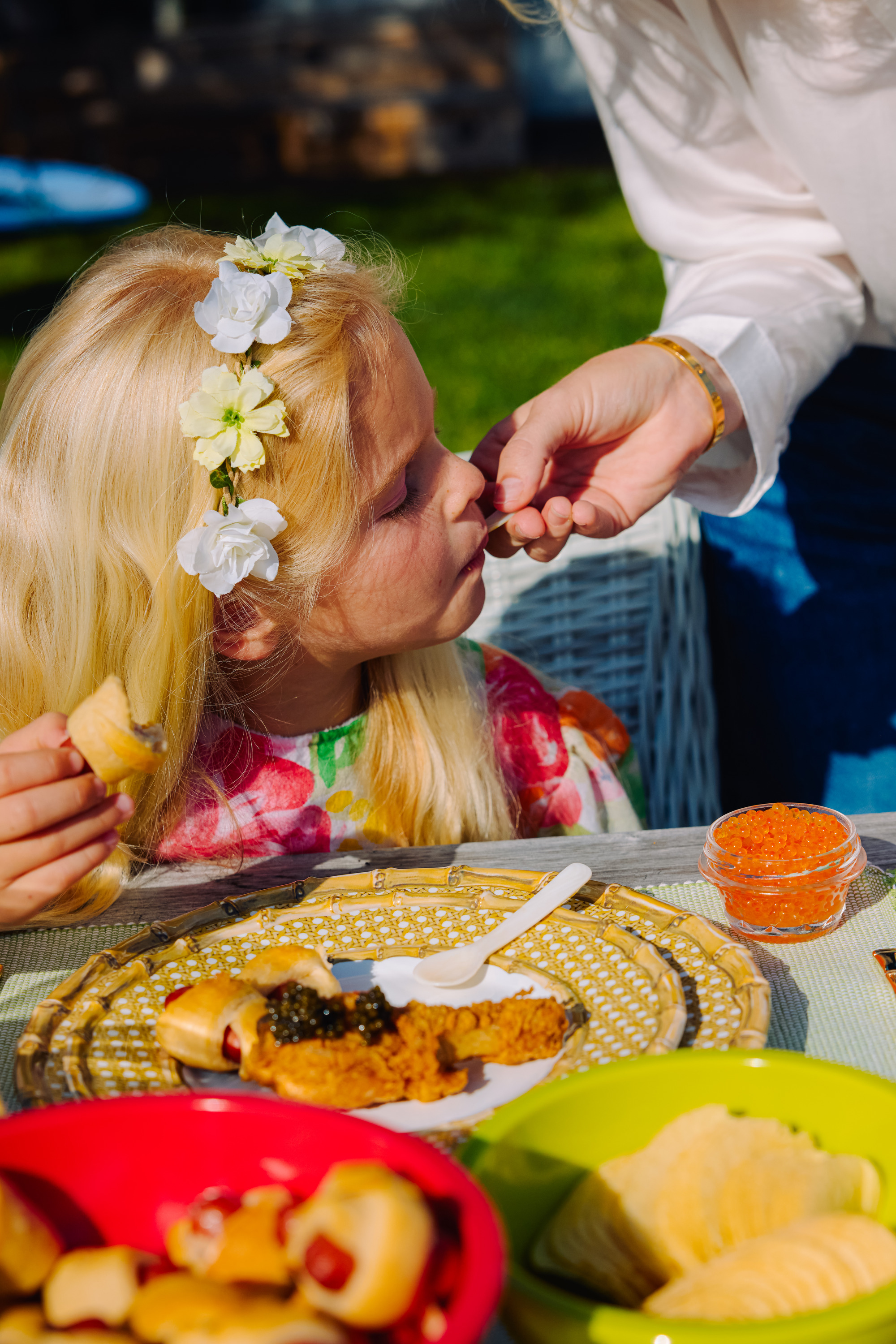 A child with a flower headband is being fed by someone in a white shirt. She's holding a small sandwich, and various snacks are on the table.
