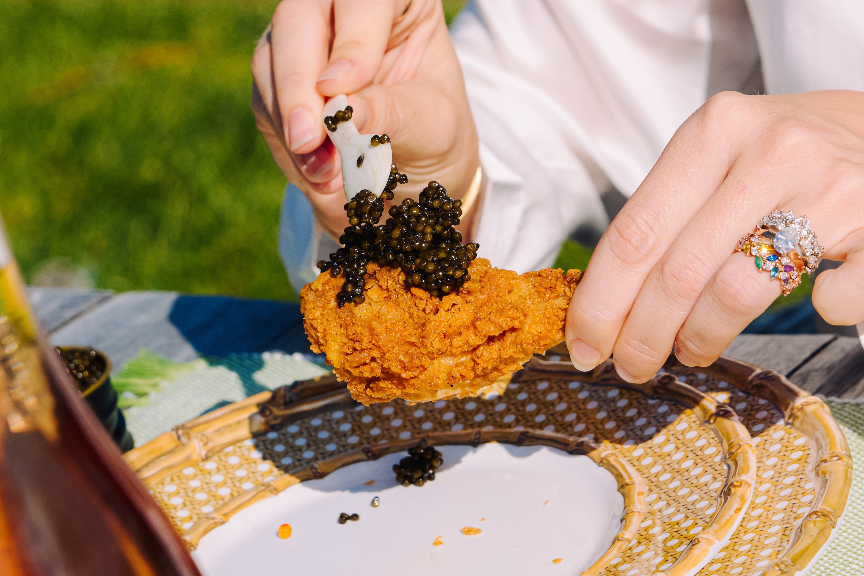 A person is placing black caviar on a piece of fried chicken using a small spoon, set on a decorative plate outdoors, with a bottle nearby.