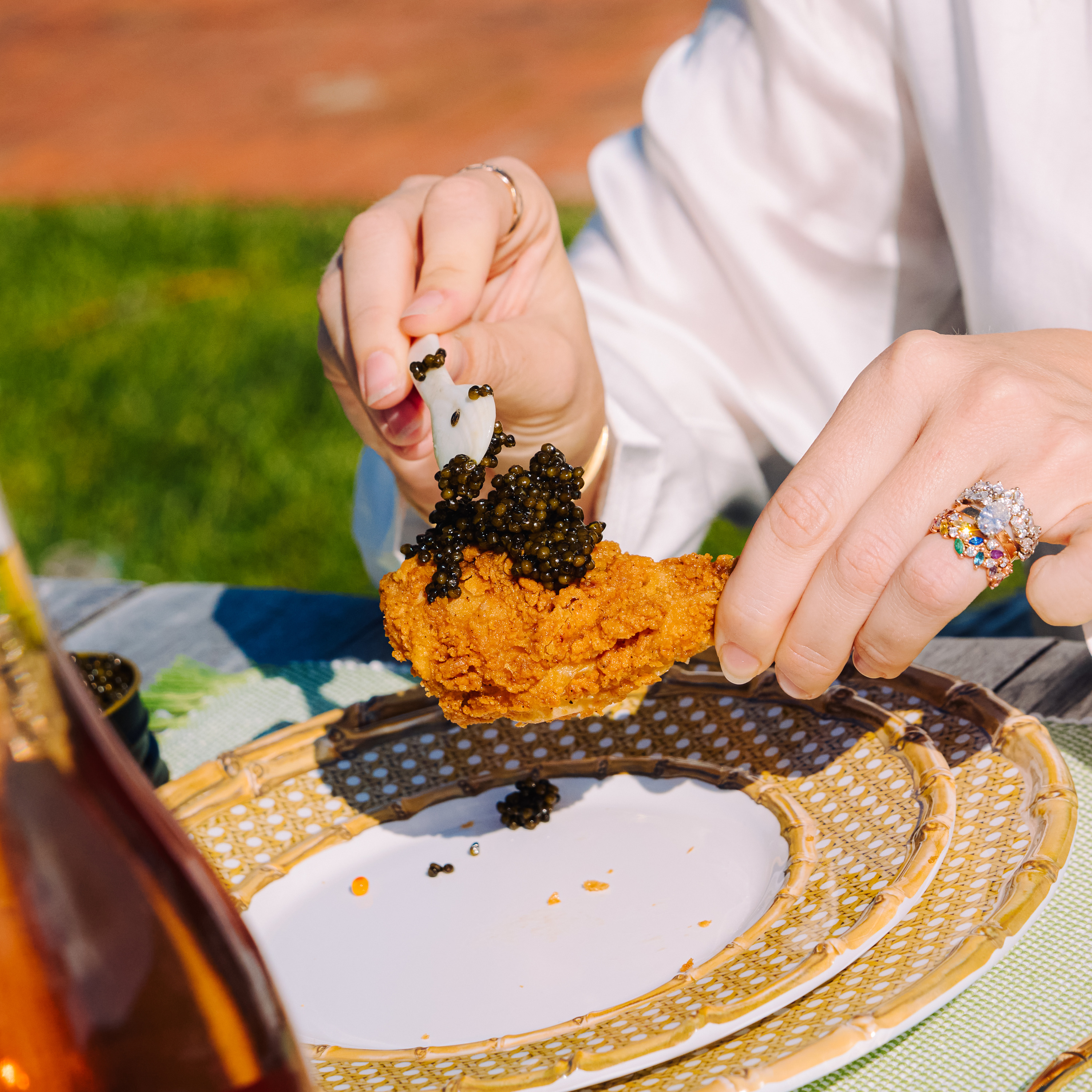 A person is placing black caviar on a piece of fried chicken using a small spoon, set on a decorative plate outdoors, with a bottle nearby.