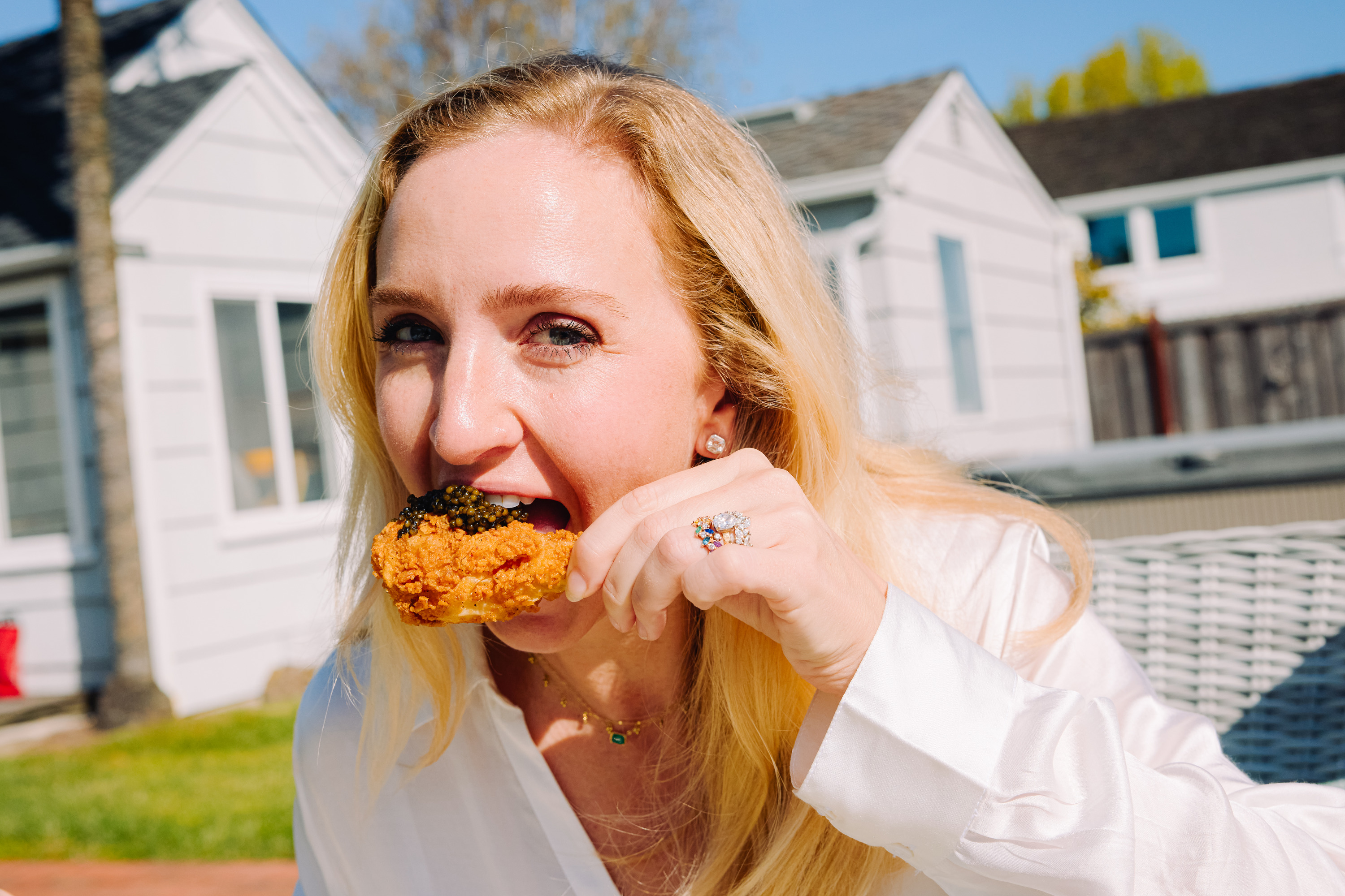 A woman with blonde hair is outdoors, eating a piece of fried chicken topped with what appears to be caviar. She is wearing a white shirt and has a colorful ring.