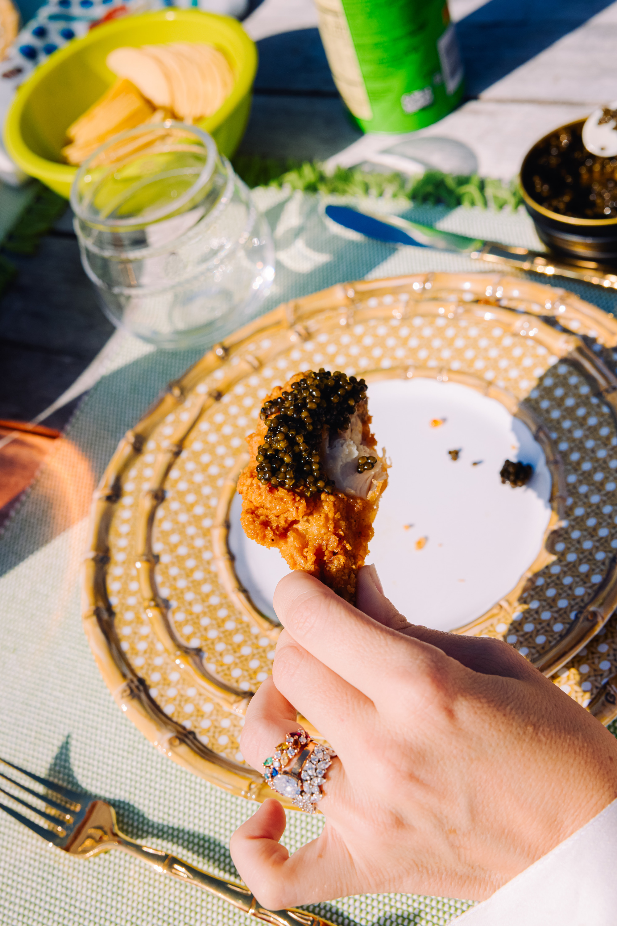 A hand with rings holds a fried chicken piece topped with caviar above a decorative plate. A bowl of snacks, a glass, and a green can are nearby on the table.