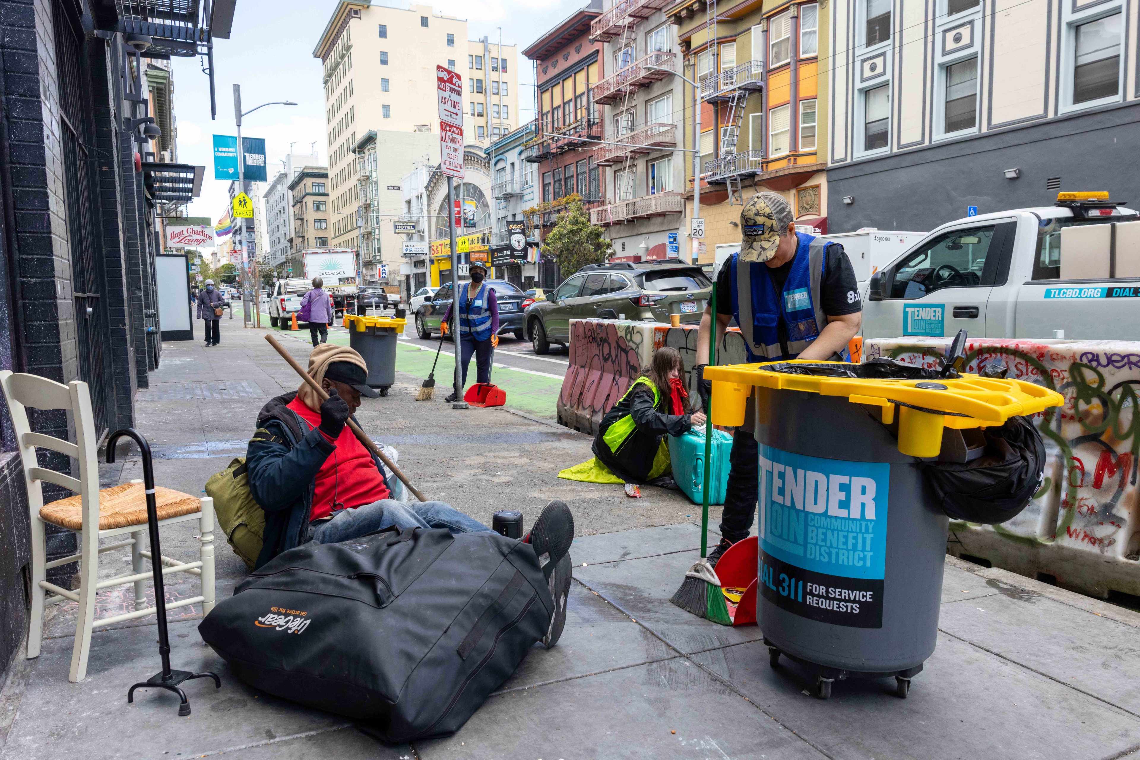 People on a city sidewalk are engaged in cleaning and organizing. One person sits with belongings, while others clean with brooms and garbage bins.