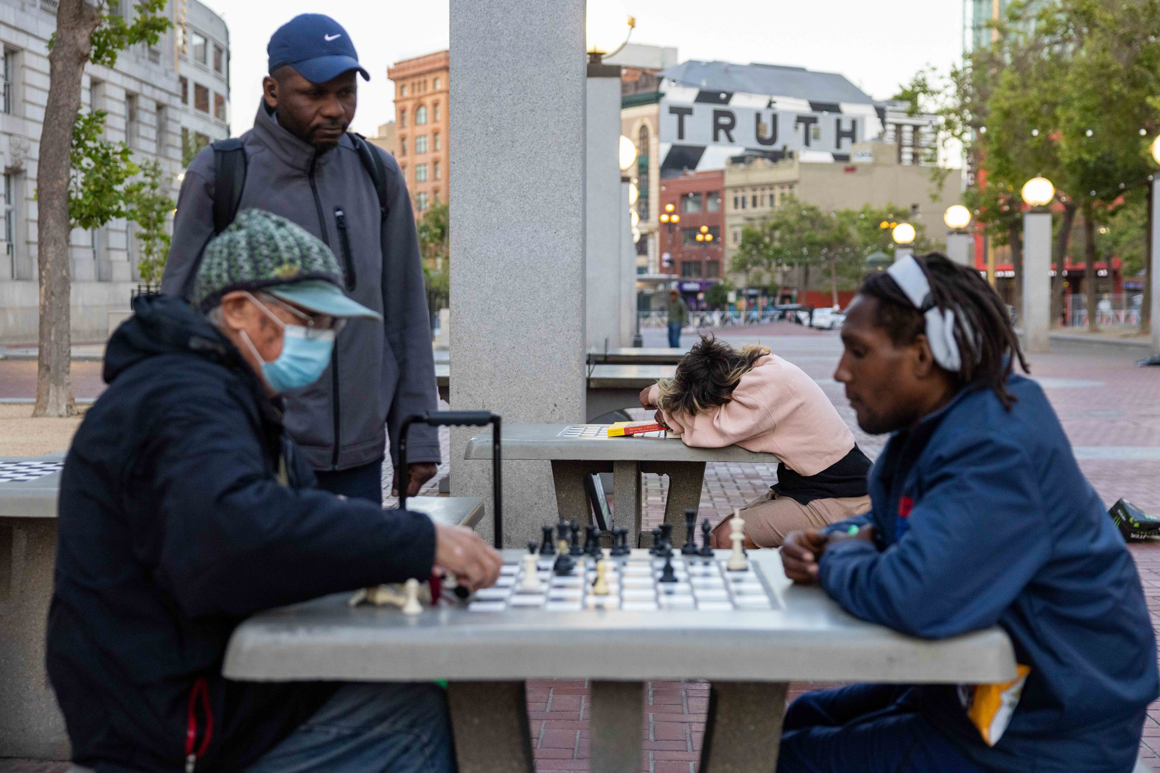 Two people are playing chess at an outdoor table, with one wearing a mask. A person stands nearby, while another rests their head on the table. The word &quot;TRUTH&quot; is visible in the background.