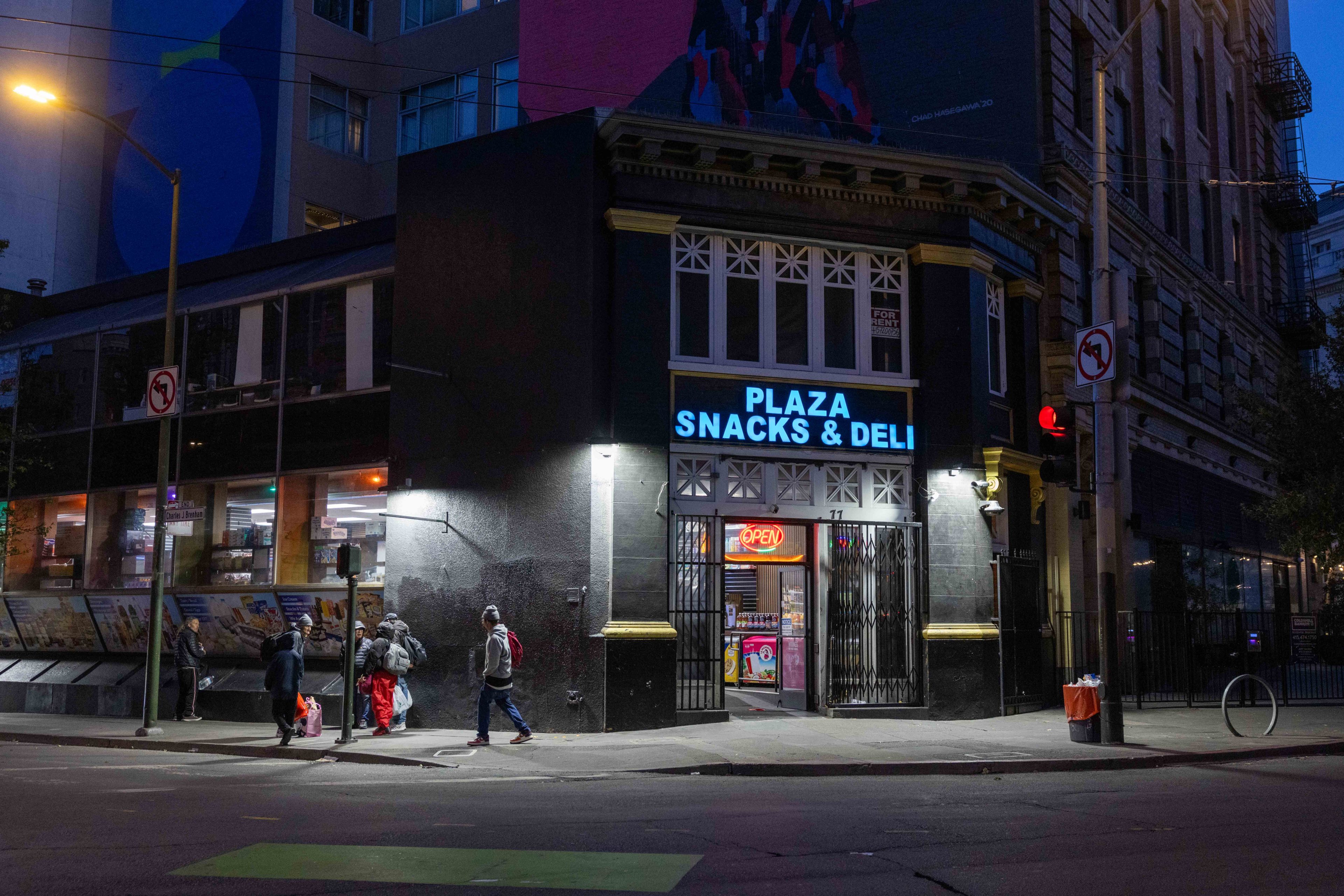 A dimly lit corner store named &quot;Plaza Snacks &amp; Deli&quot; with an open sign emits a warm glow. People with bags walk past on the sidewalk, and a no-left-turn sign is visible.