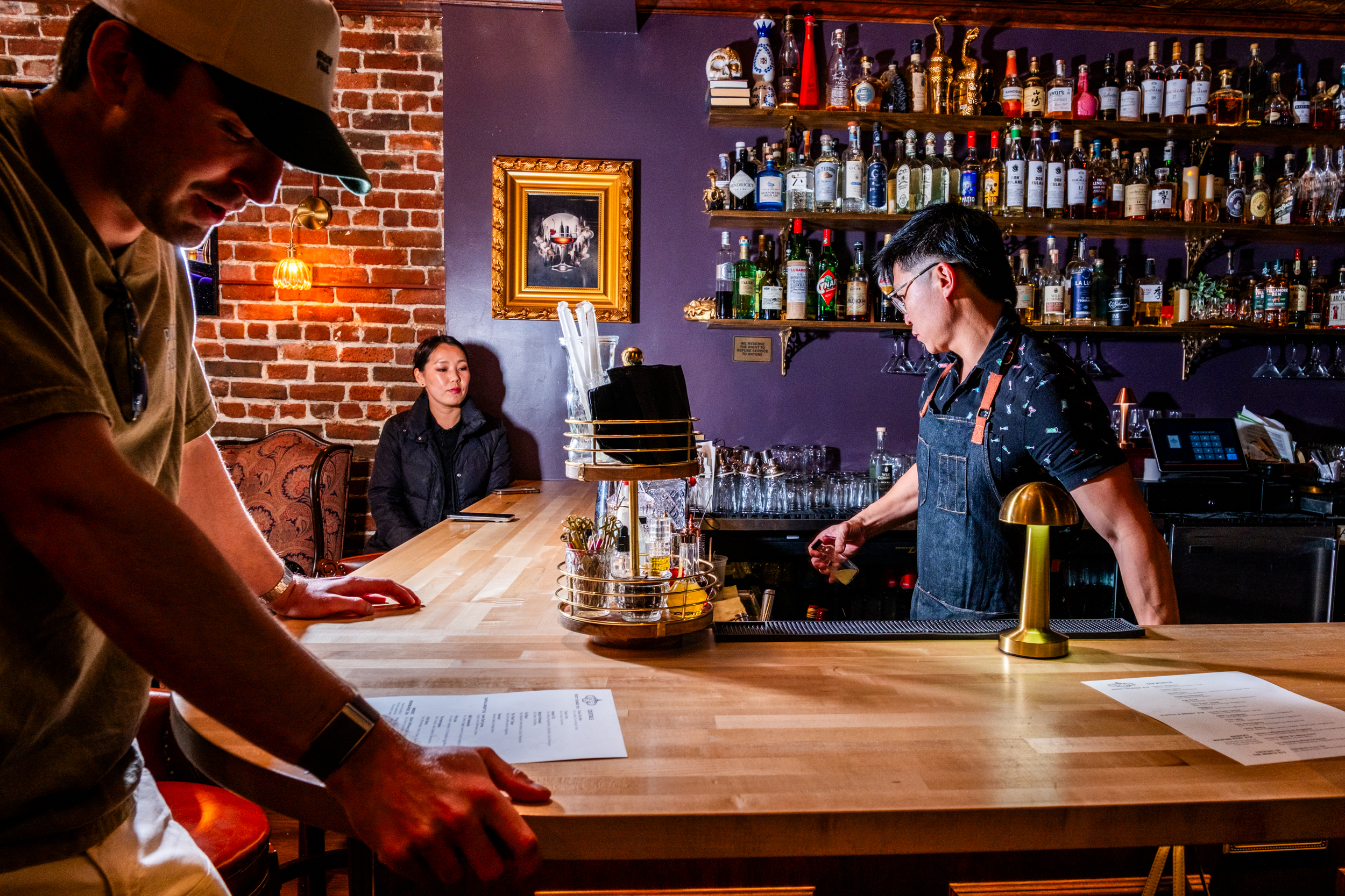 A cozy bar scene with a bartender serving drinks. A man leans on the counter, and a woman sits by the brick wall. Shelves full of bottles are in the background.