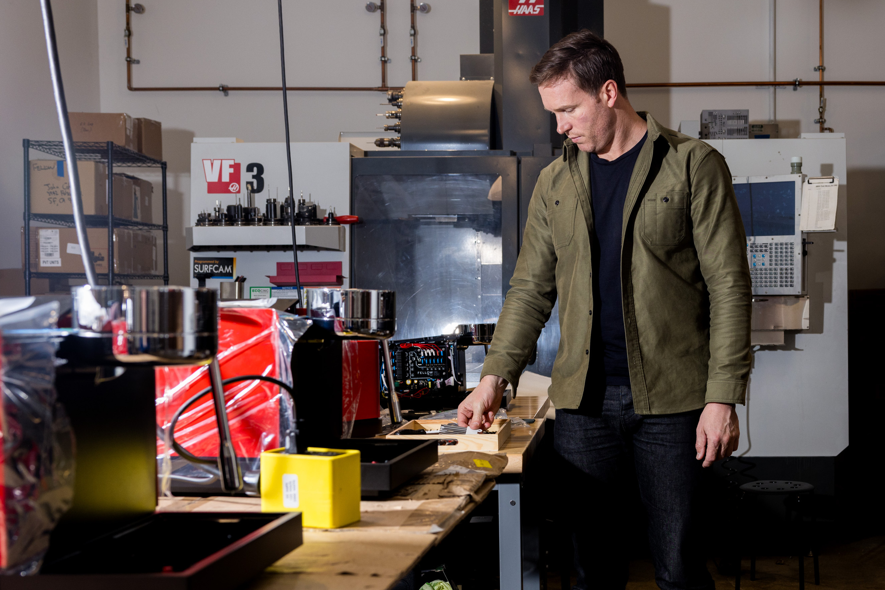A man in a green jacket stands at a workbench in a workshop, examining items. Behind him are industrial machines and shelves with tools and items.