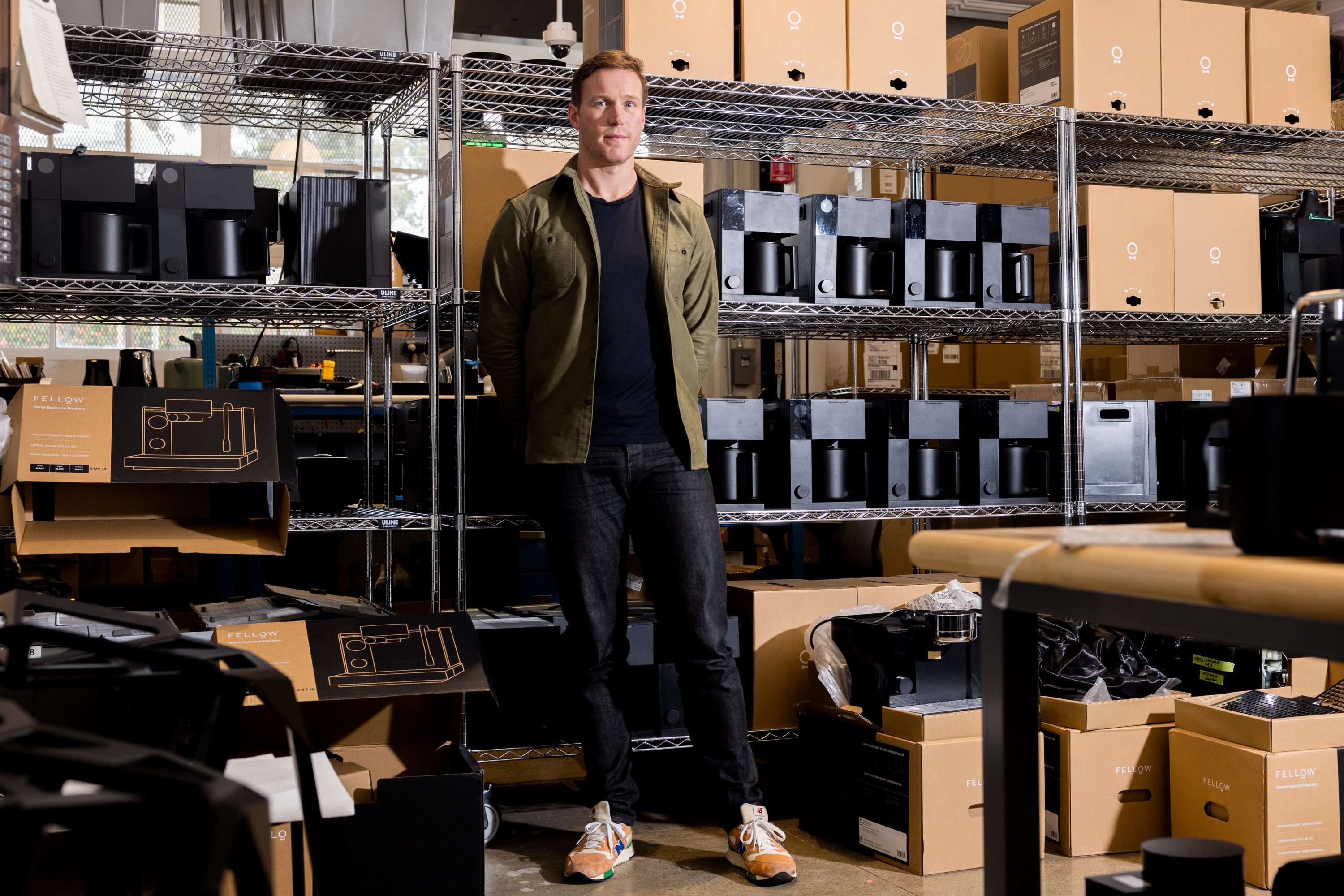 A person stands in a warehouse, surrounded by metal shelves filled with black and brown boxes. Some boxes are open, showing products inside. The floor is concrete.