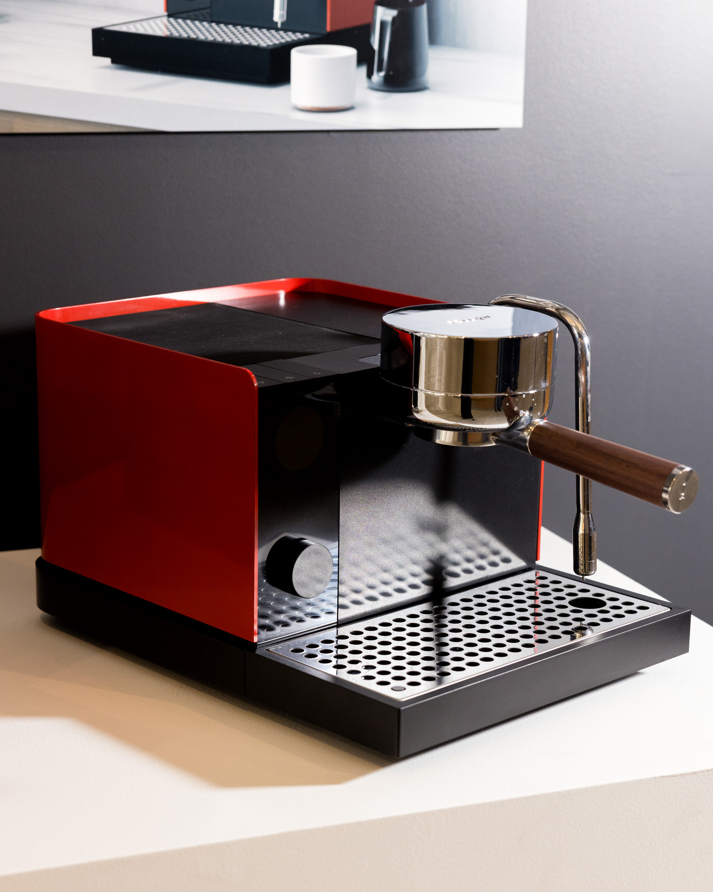 A sleek red espresso machine sits on a counter with a chrome portafilter and a textured black drip tray. A similar machine is shown in the background poster.