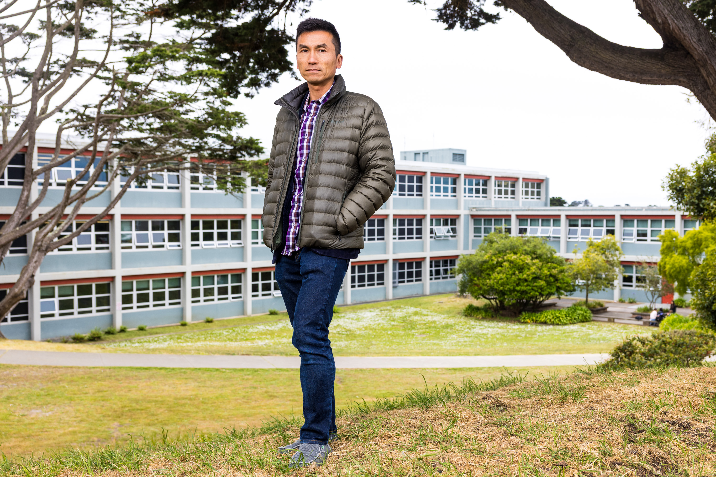 A person in a plaid shirt and puffy jacket stands on grassy ground near a tree. Behind them is a modern building with large windows and a lawn with bushes.