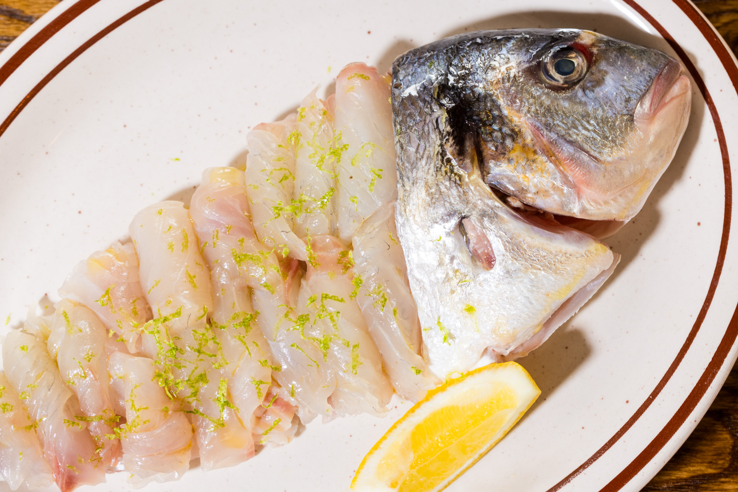 The image shows a fish head with sliced raw fish fillets arranged on a plate, garnished with green zest and a lemon wedge beside it.