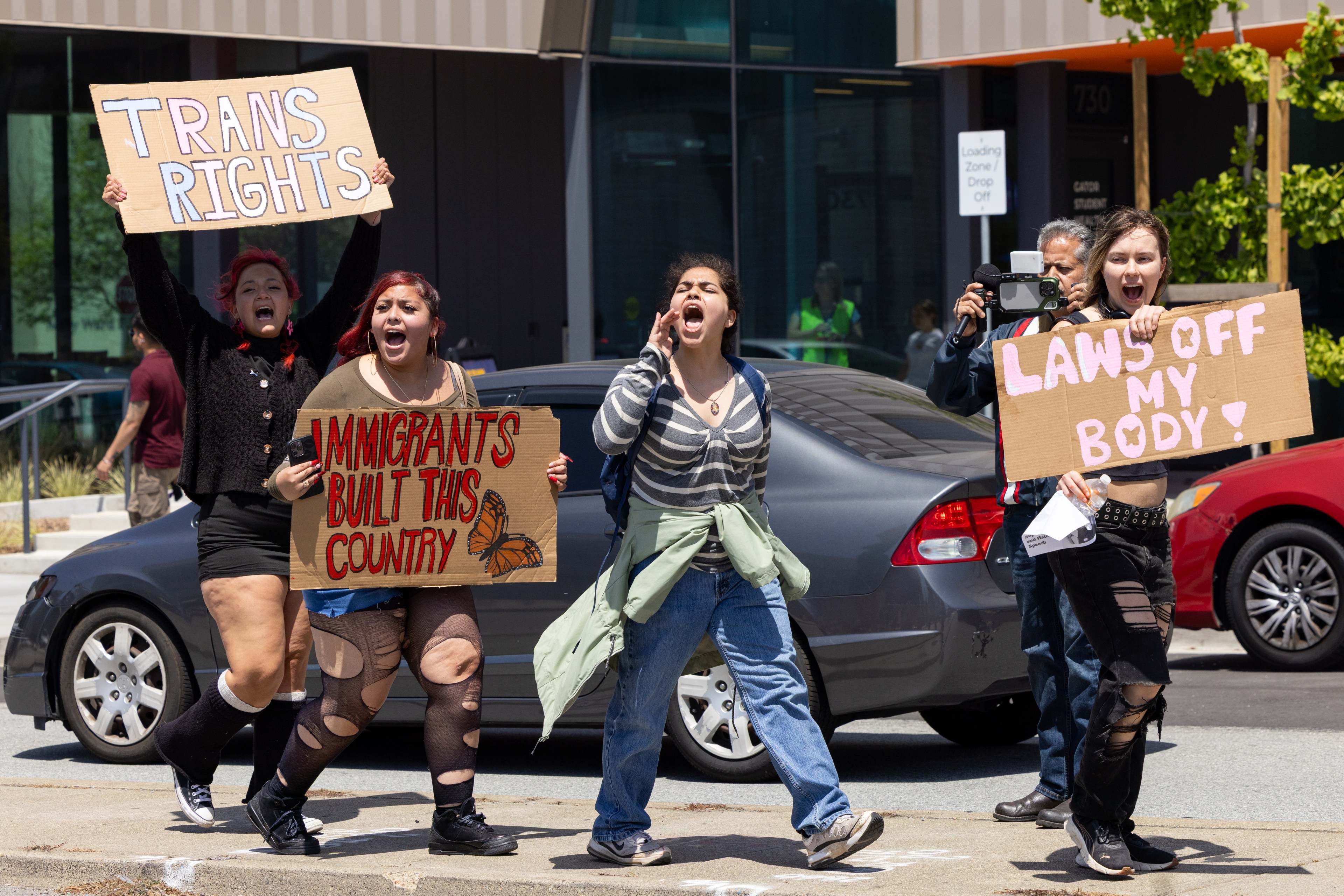 A group of people protest energetically on a street, holding signs advocating for trans rights, immigrant contributions, and bodily autonomy.