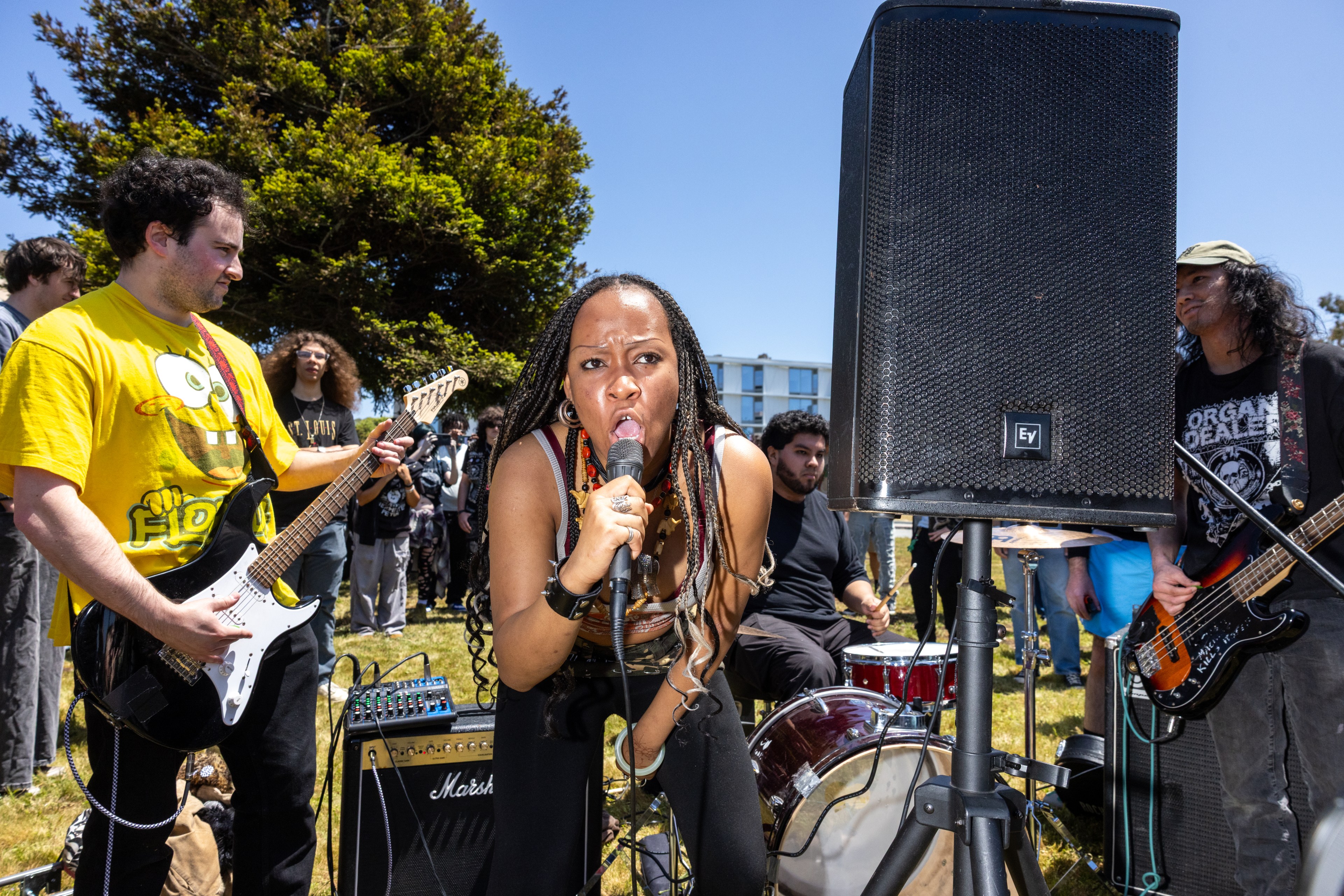 A dynamic outdoor band performance is shown with a vocalist passionately singing into a microphone, flanked by musicians playing guitar and drums under a clear blue sky.