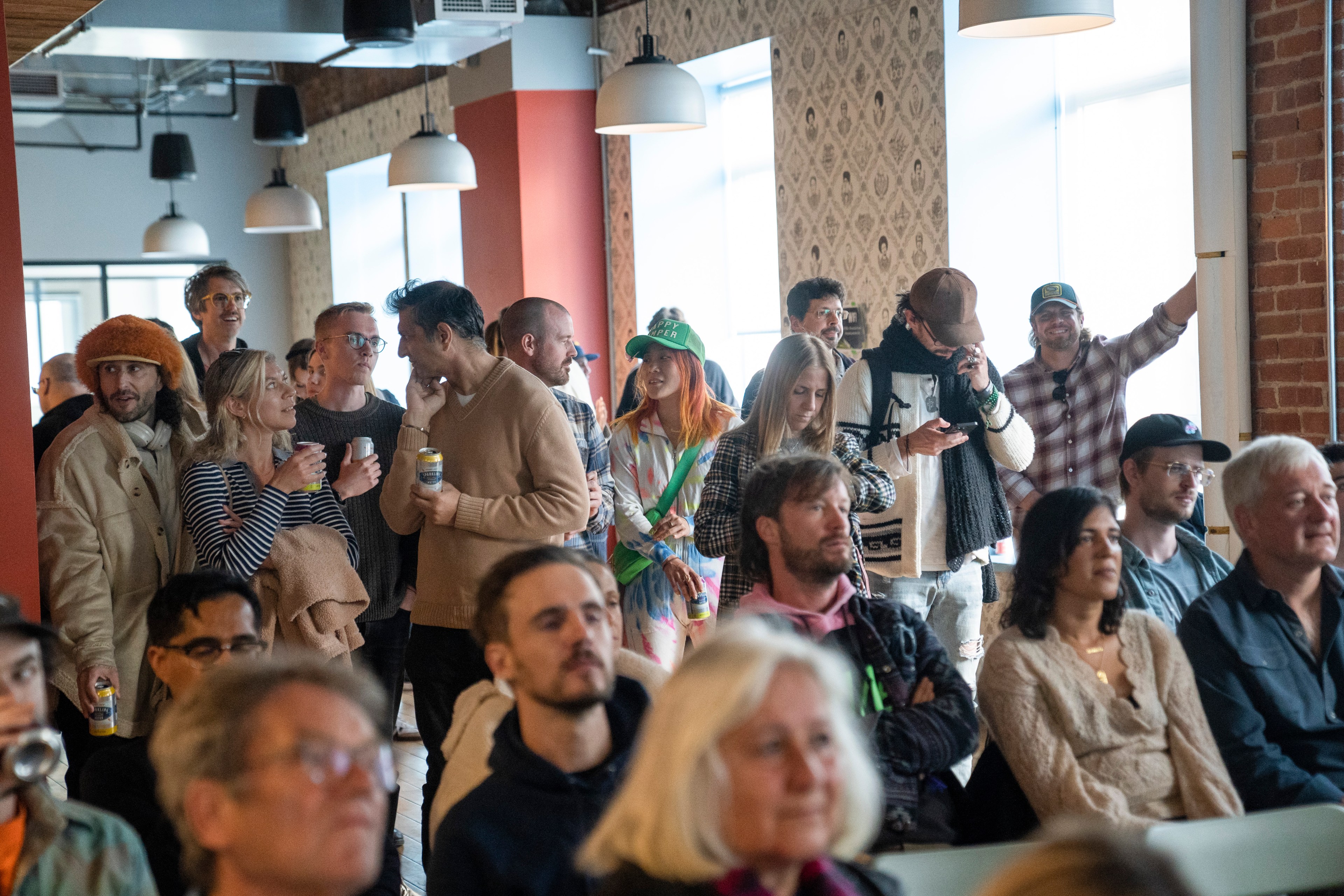 A group of people gathers in a well-lit room, some standing and chatting with drinks, others seated attentively, all under modern hanging lights.