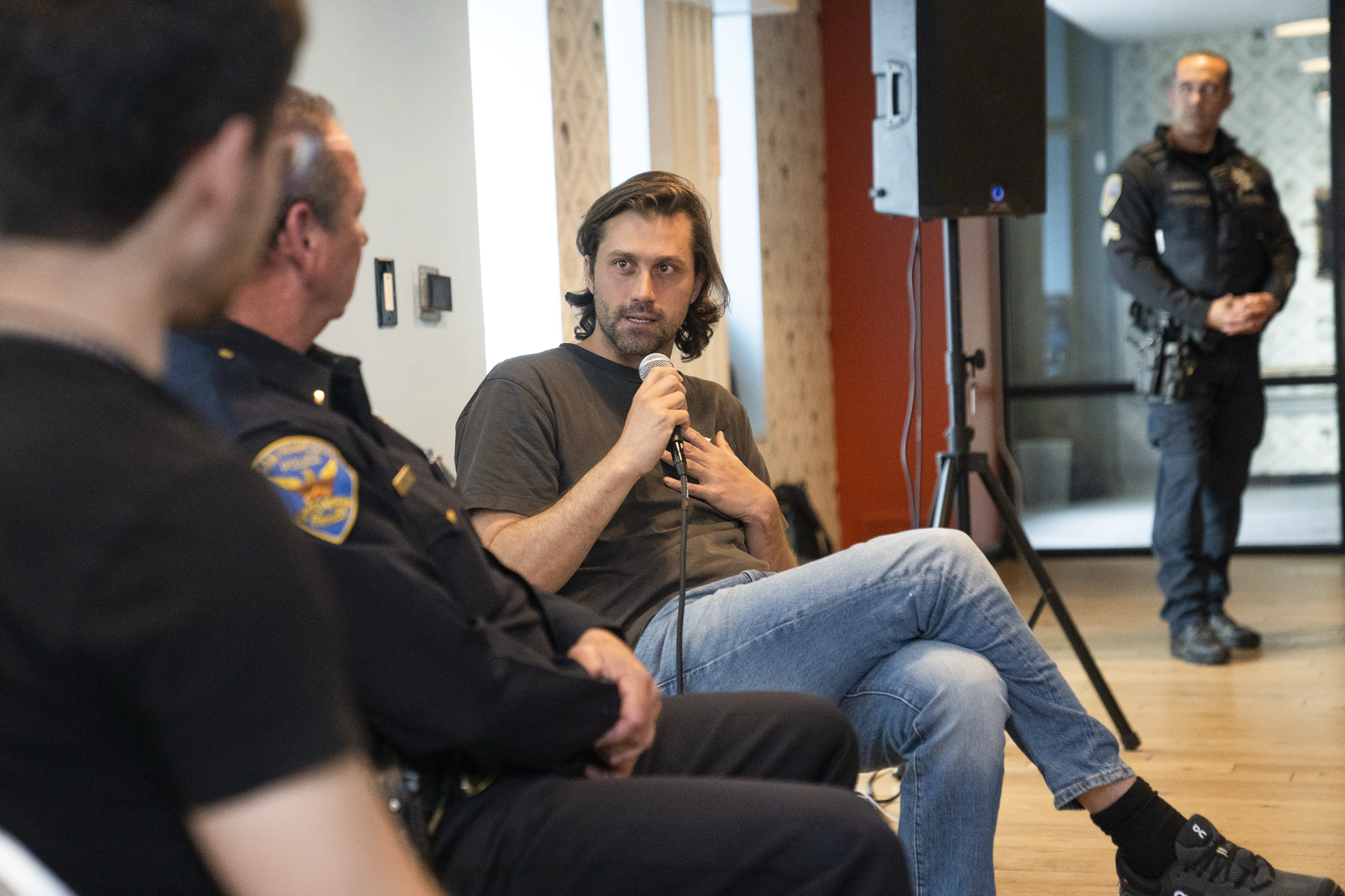 A man in casual clothes speaks into a microphone while sitting with two uniformed officers. Another officer stands in the background near a speaker.