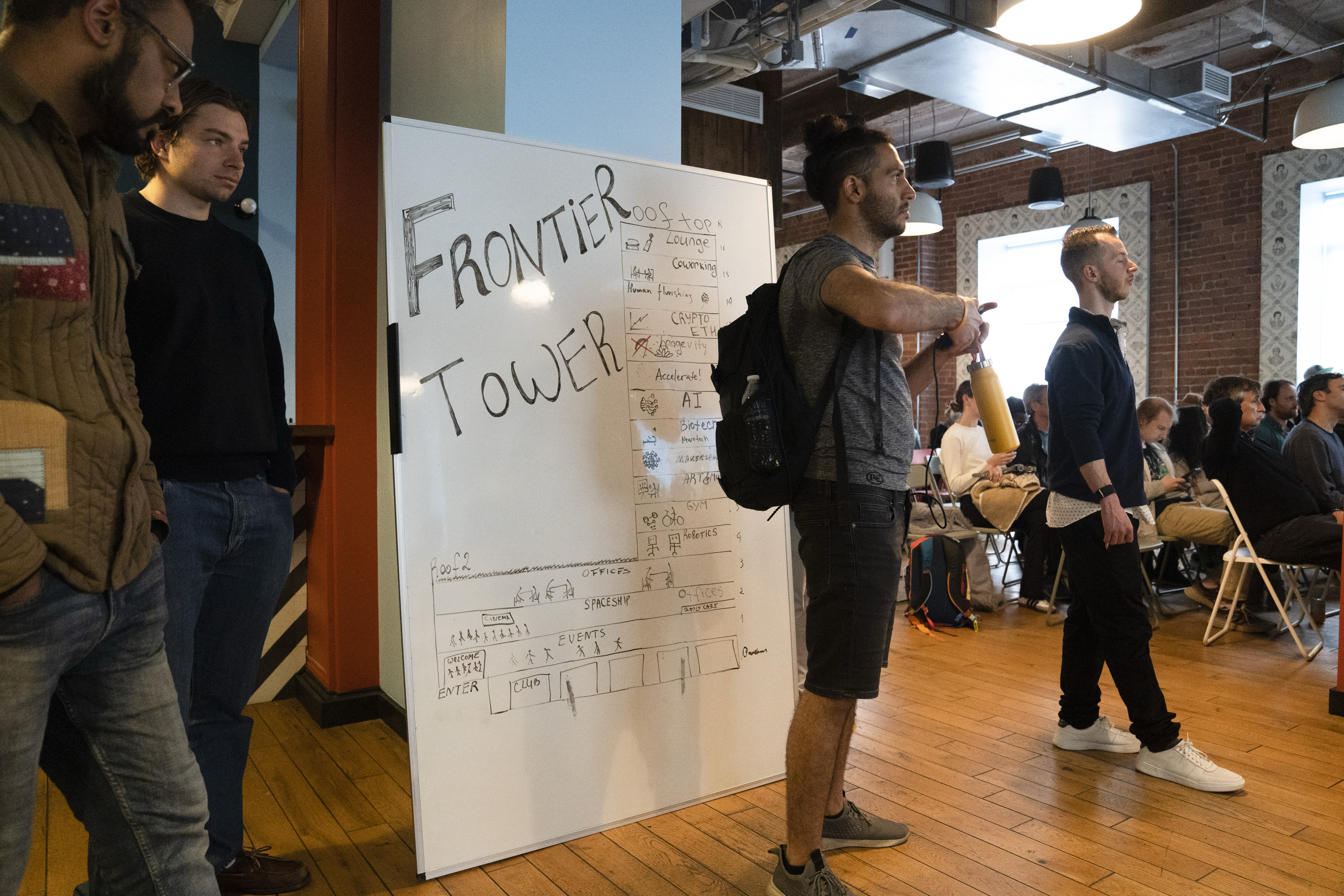 A group of people stand near a whiteboard with "FRONTIER TOWER" written on it. The room is filled with seated attendees and has a wooden floor and brick walls.