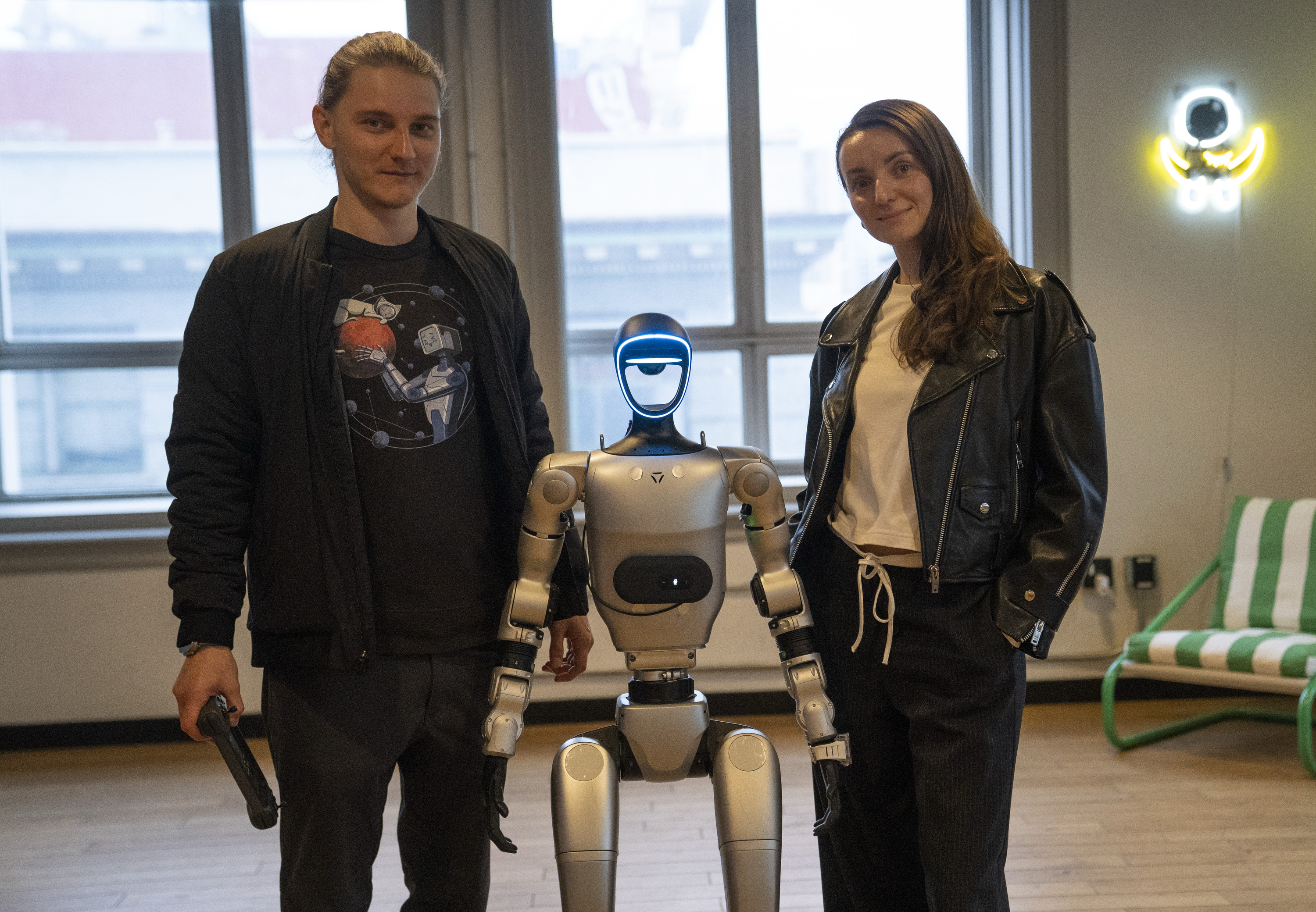 A man and a woman stand on either side of a humanoid robot with a sleek design. The room has large windows in the background and a neon wall decoration.