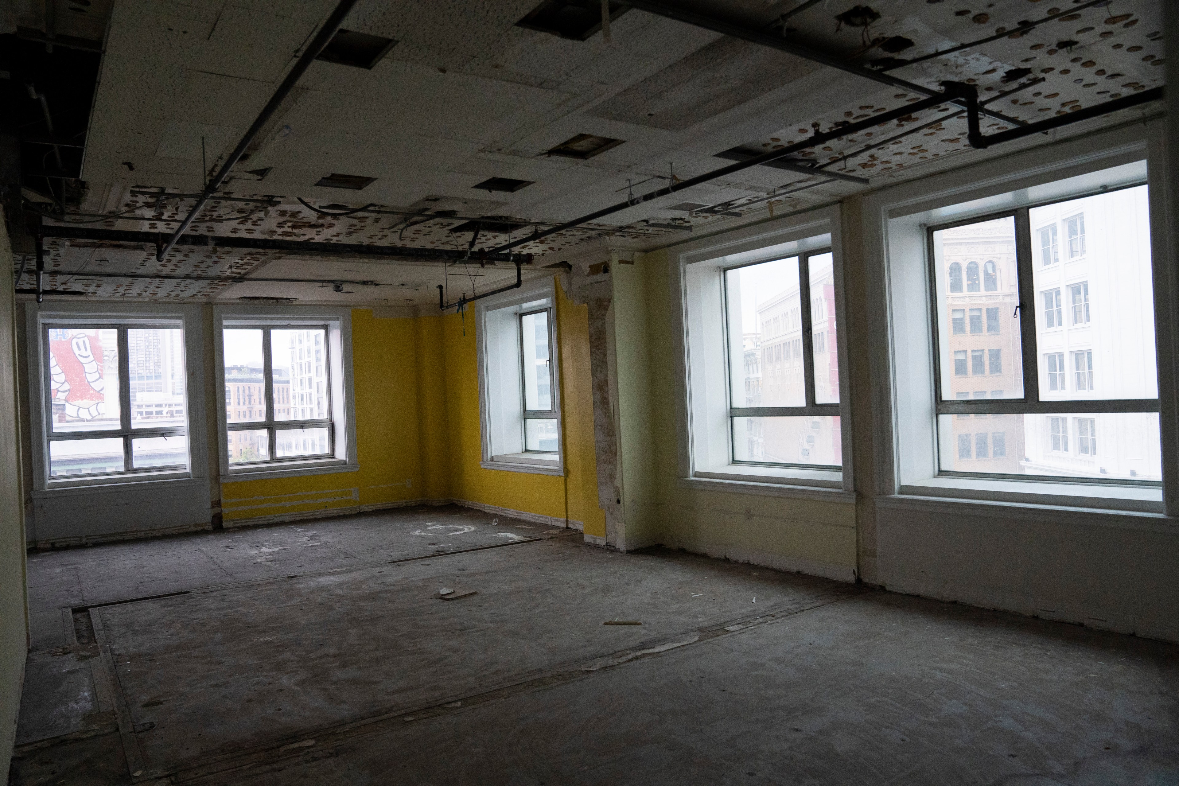 An empty room with stripped floors and patches on the ceiling. Three large windows let in natural light, and the walls are painted a faded yellow.