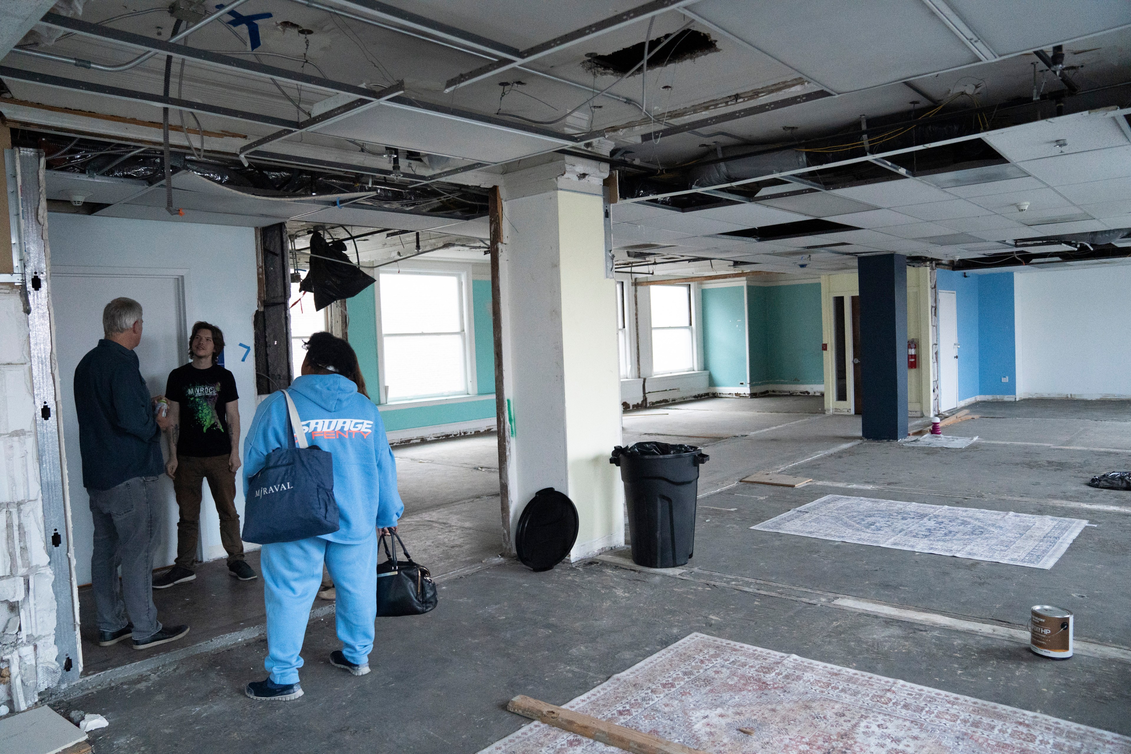 Three people stand in a partially demolished room with exposed ceilings and unfinished floors. Rugs and a trash can are on the floor, with walls painted teal and white.