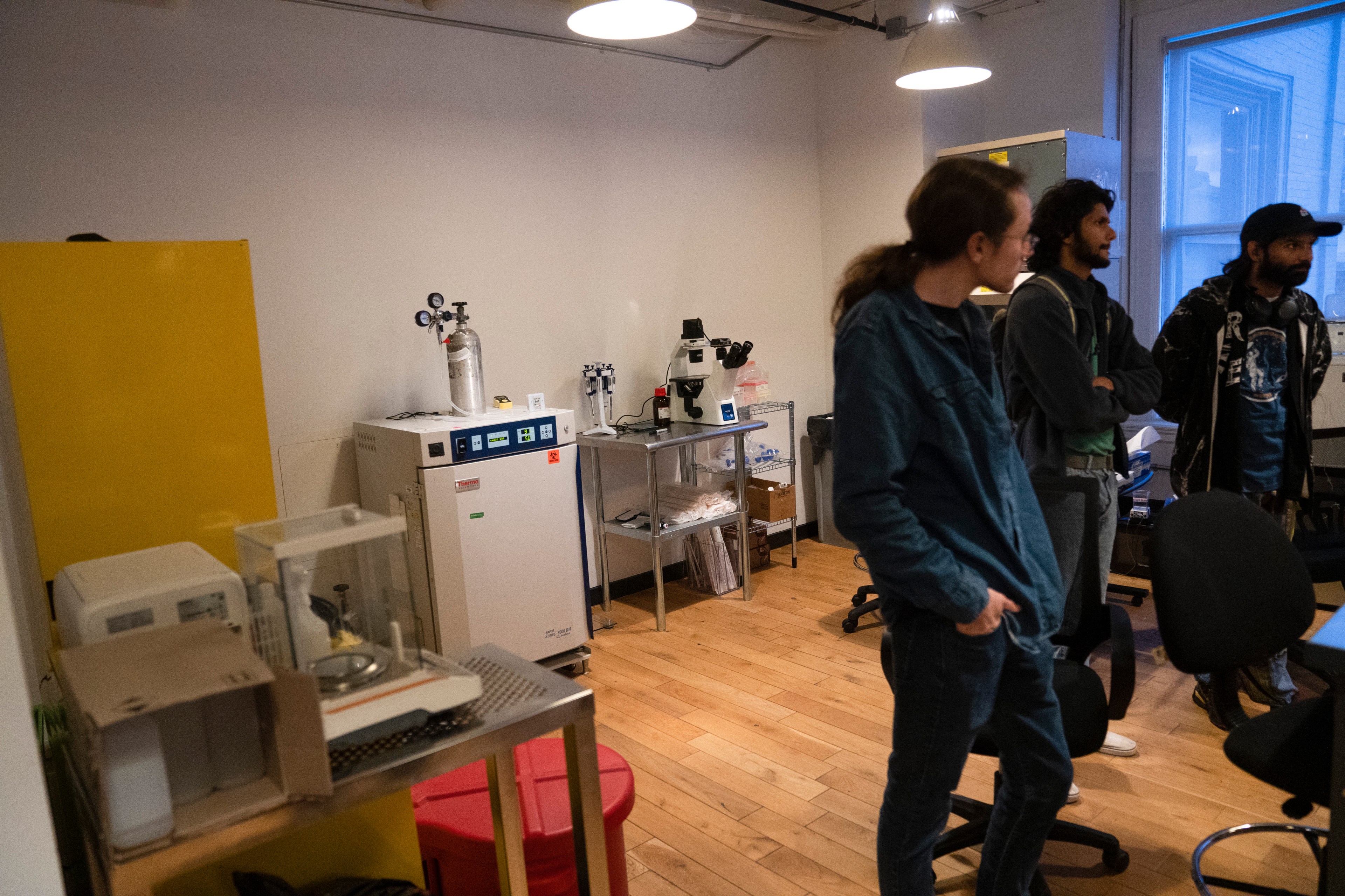 A group of people stands in a lab with a microscope, equipment, and wooden flooring. There's a yellow panel on the left and a window on the right.