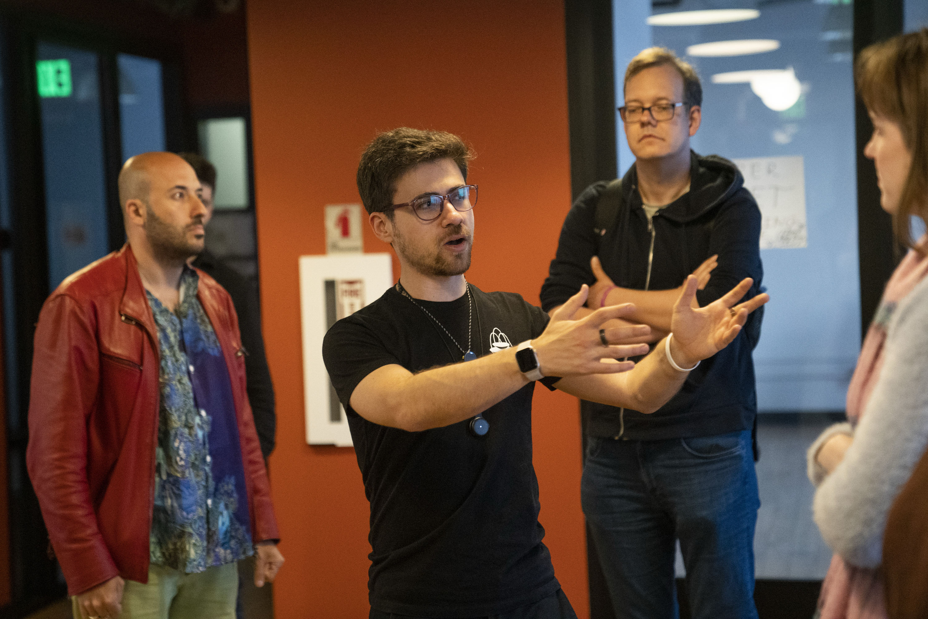 A man in glasses and a black shirt gestures while speaking to a small group of people in a room with orange walls and dim lighting.