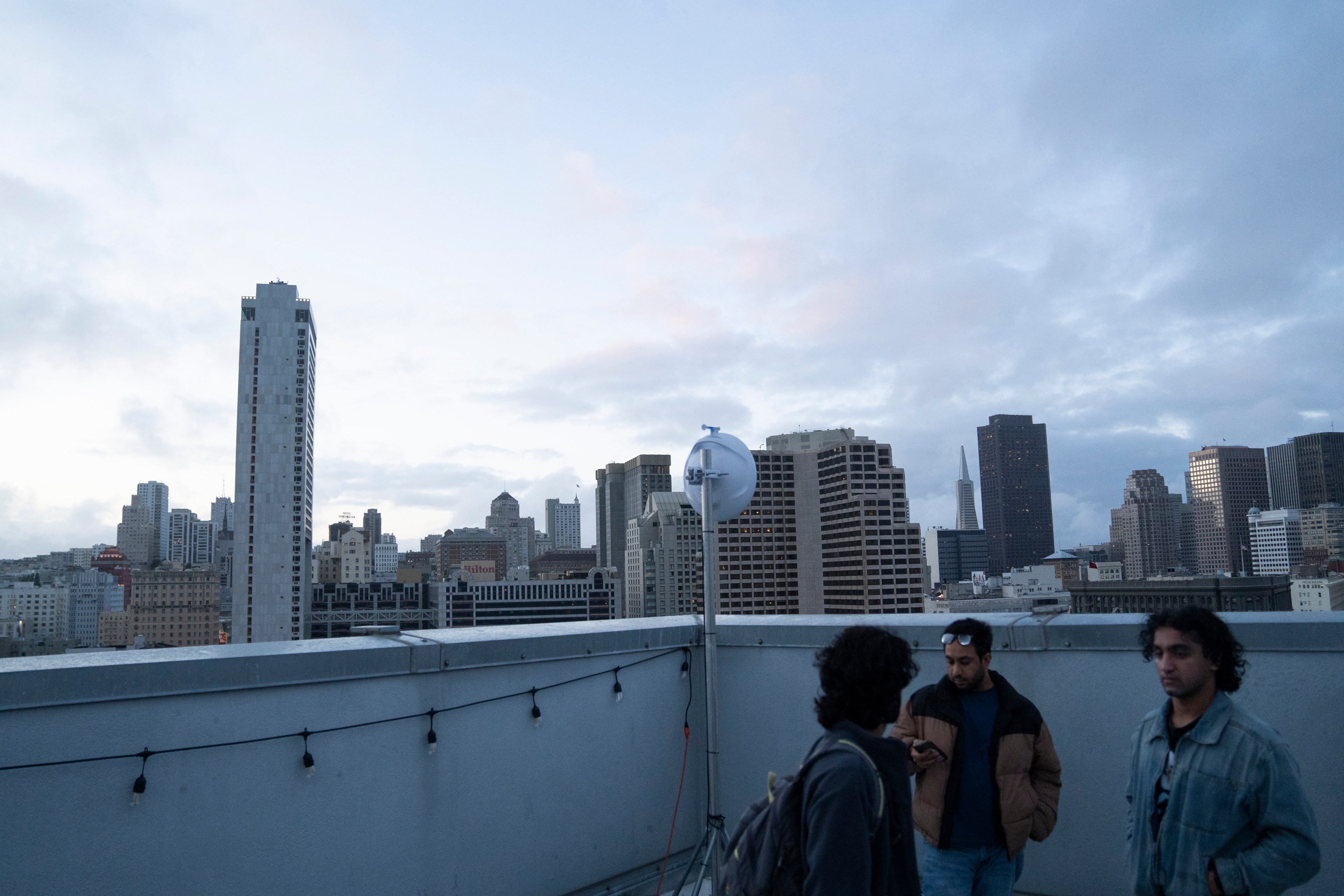 Three people are standing on a rooftop with a city skyline in the background. The sky is cloudy, and decorative lights and a satellite dish are visible.