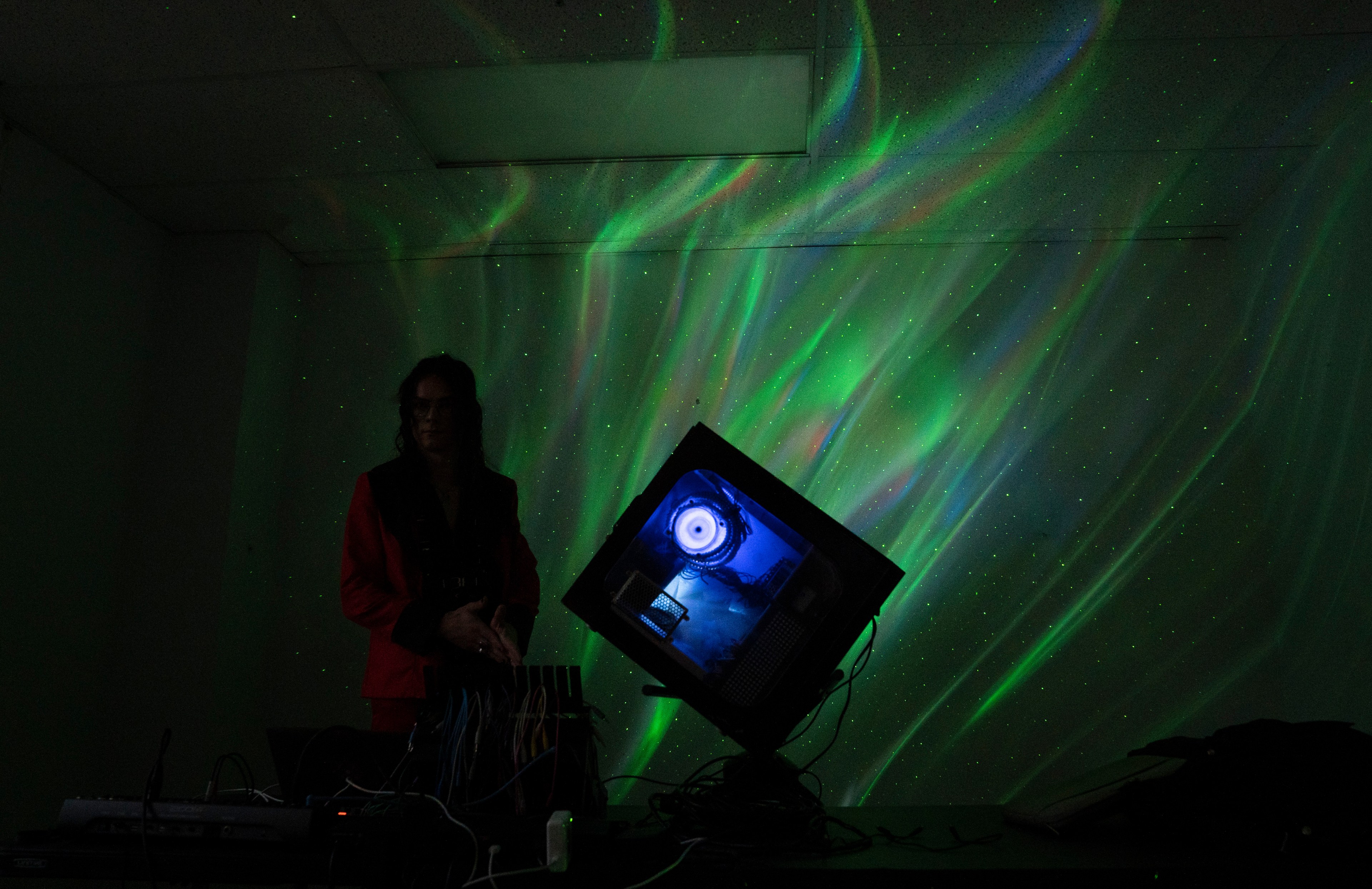 A person stands in a dark room with a colorful aurora-like light display on the wall. A tilted monitor on a table shows a circular, eye-like design.