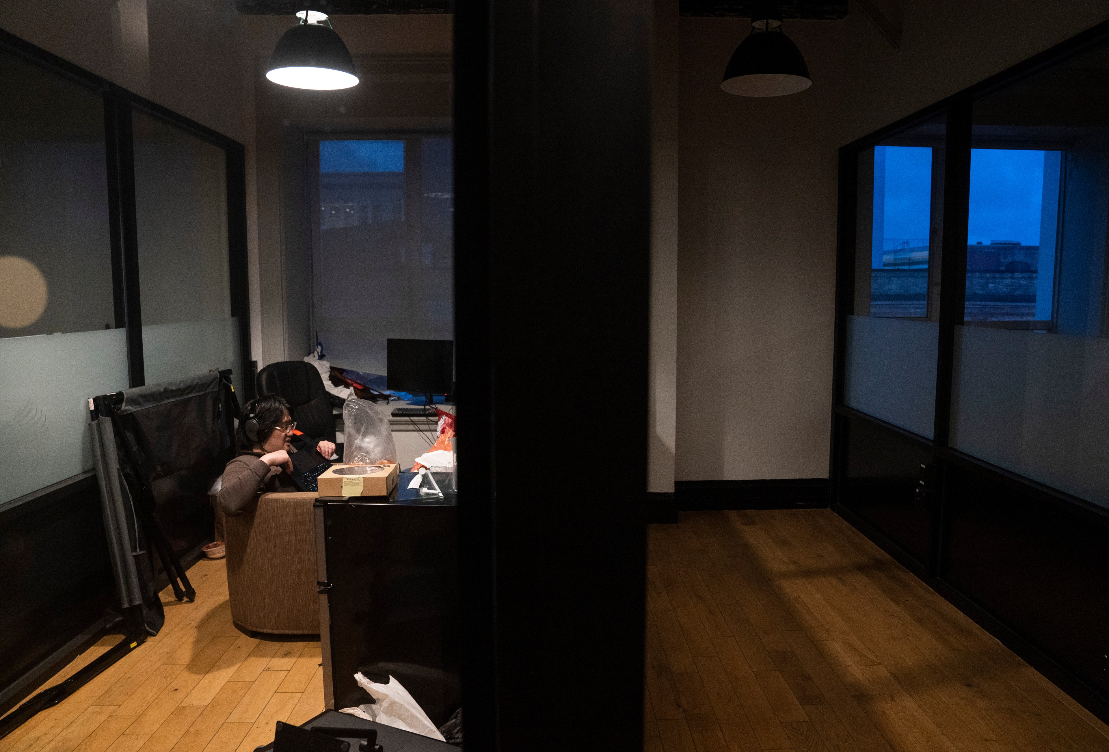 A person is sitting at a cluttered desk in a dimly lit room, wearing headphones and likely working on a computer. The room is divided by a glass wall.