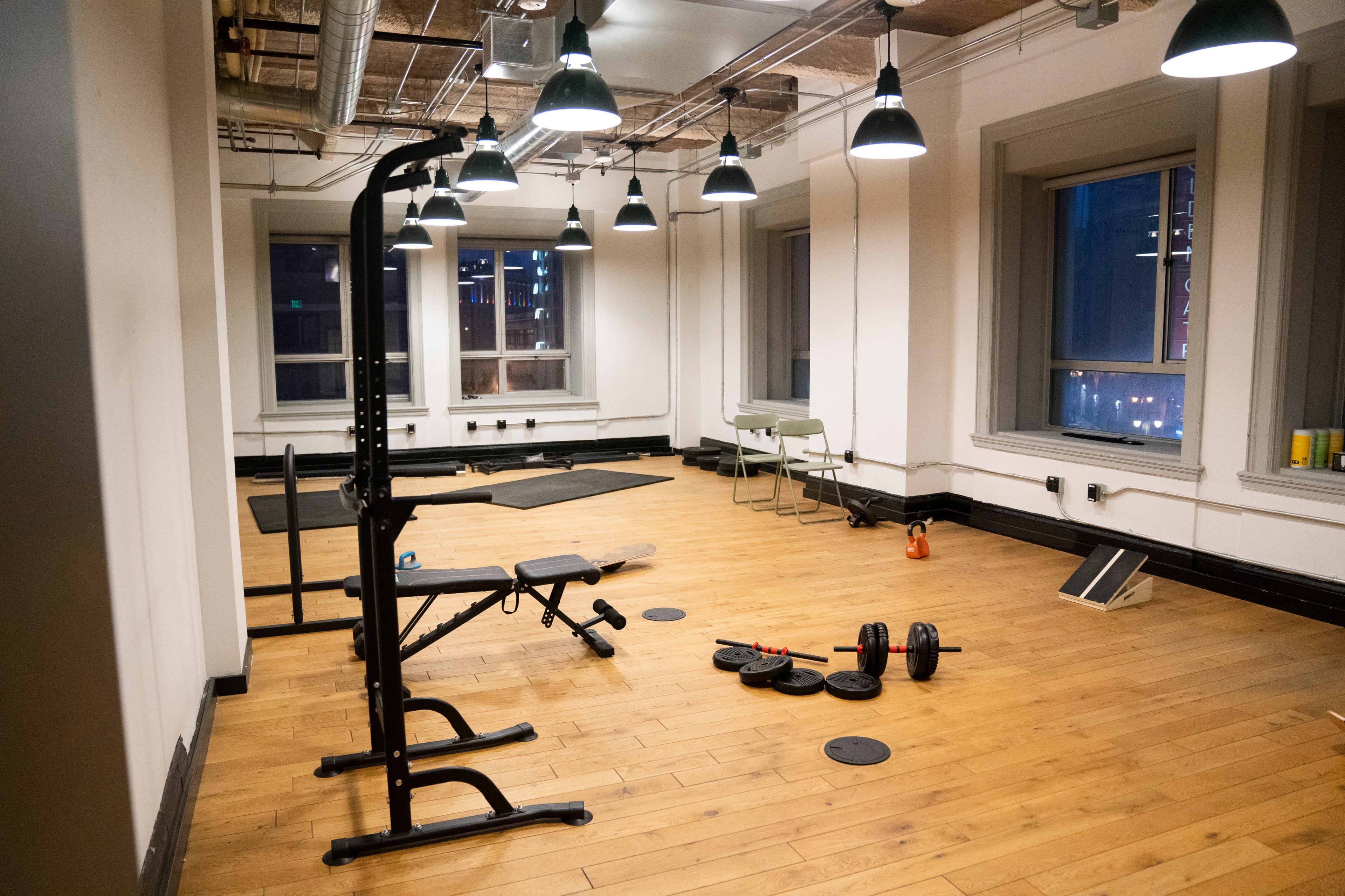 A gym room with wooden floors, featuring a weight bench, dumbbells, kettlebells, mats, a chair, and padded flooring near large windows under hanging lights.
